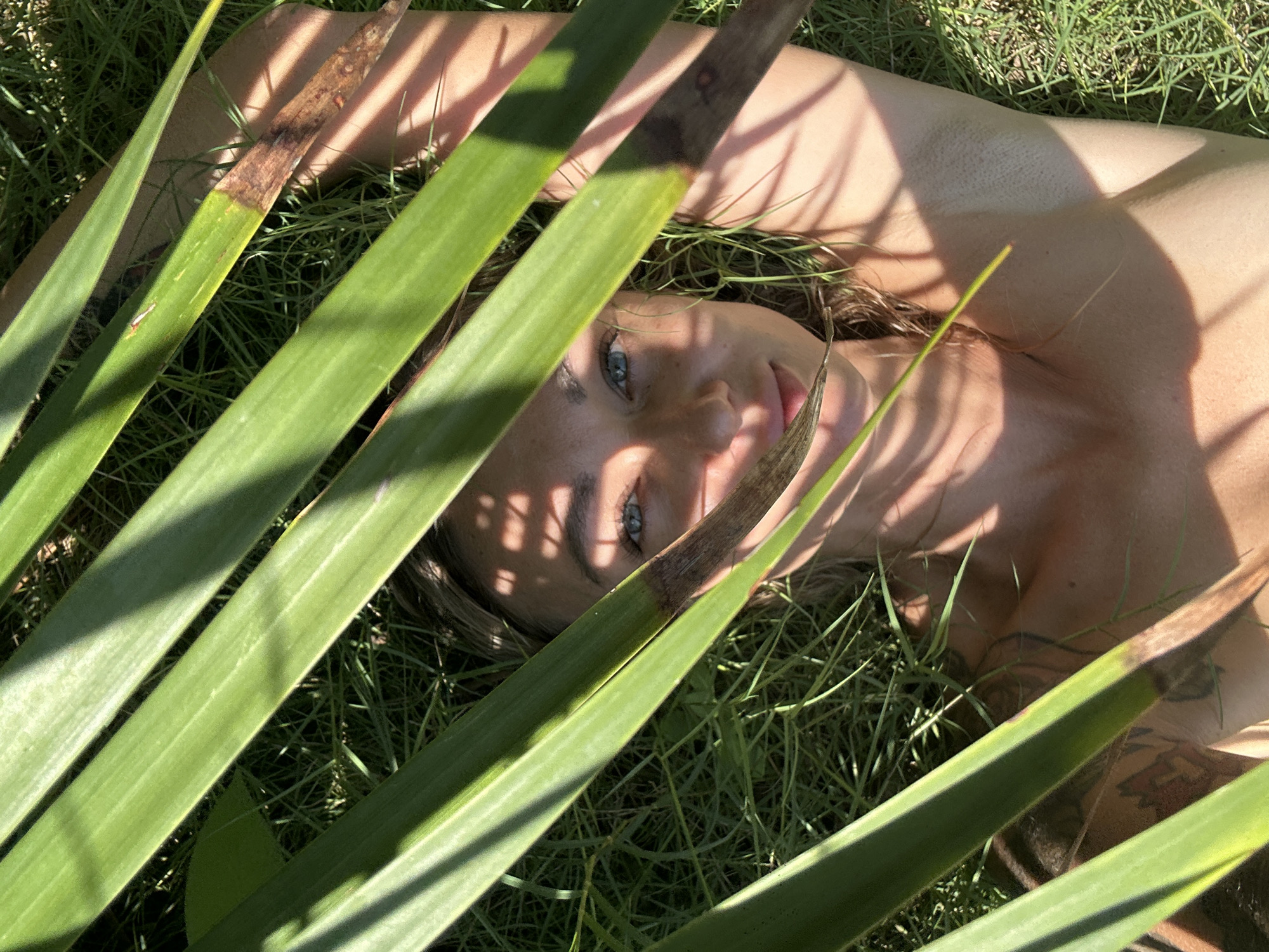 A fair-skinned person is lying on their back in a grassy area, partially obscured by long, green leaves. The person's face, including blue eyes and pink lips, is visible. Shadows cast by the leaves fall across the person's face and body. The person has tattoos on their body. The setting appears to be a natural environment, likely in Mũi Né, Vietnam, based on the presence of lush vegetation.