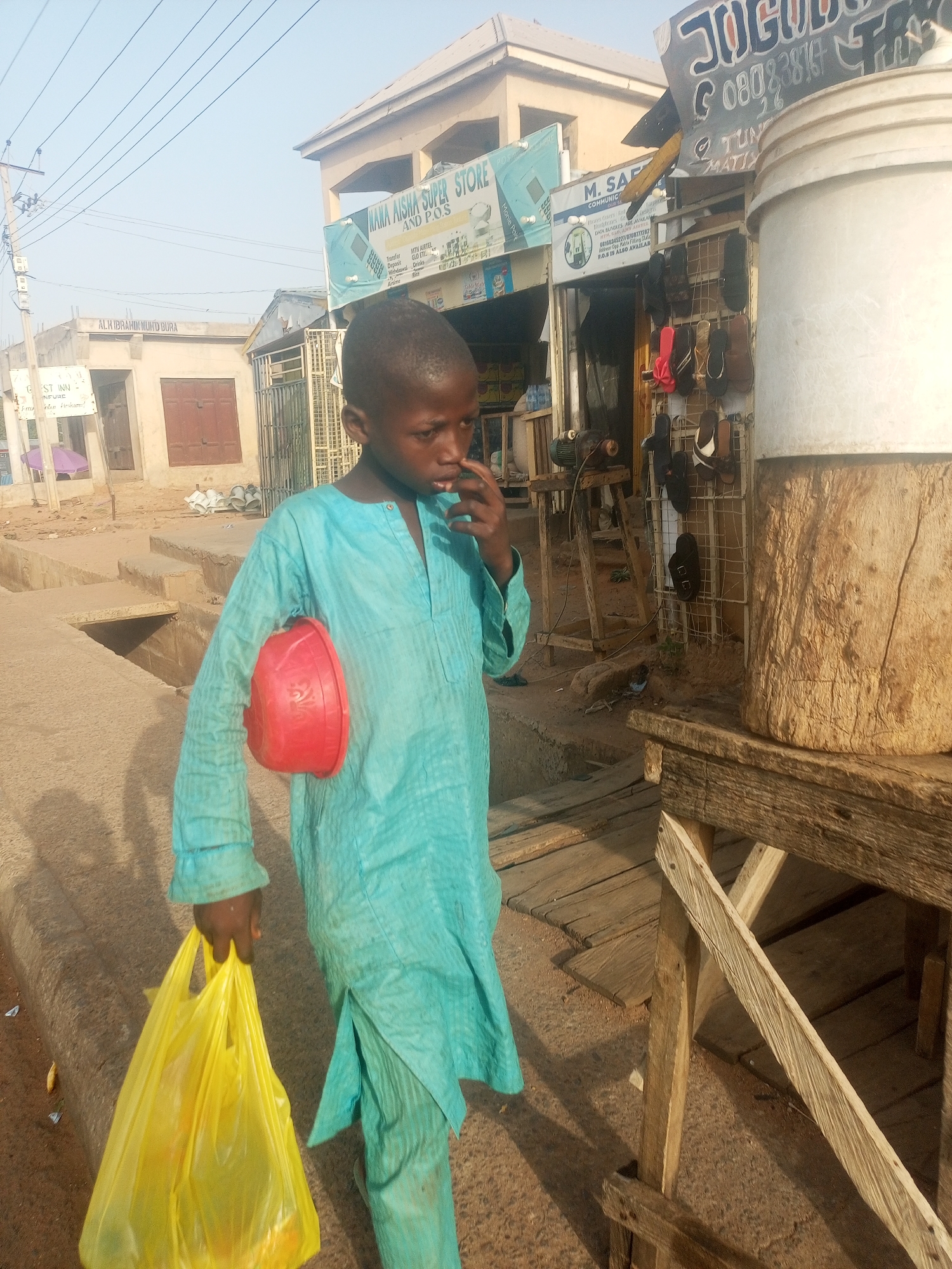 A young boy in a light blue tunic stands on a dusty street in Garko, Nigeria. He holds a yellow plastic shopping bag in his left hand and a red plastic bowl under his left arm, while his right hand is near his face. Behind him is a roadside vendor's stall with sandals displayed for sale, and a white bucket on a wooden stand. Signs for local businesses like "Nama Aisha Super Store" and "M. Saf Communication" are visible in the background. The area appears to be a mix of residential and commercial buildings.