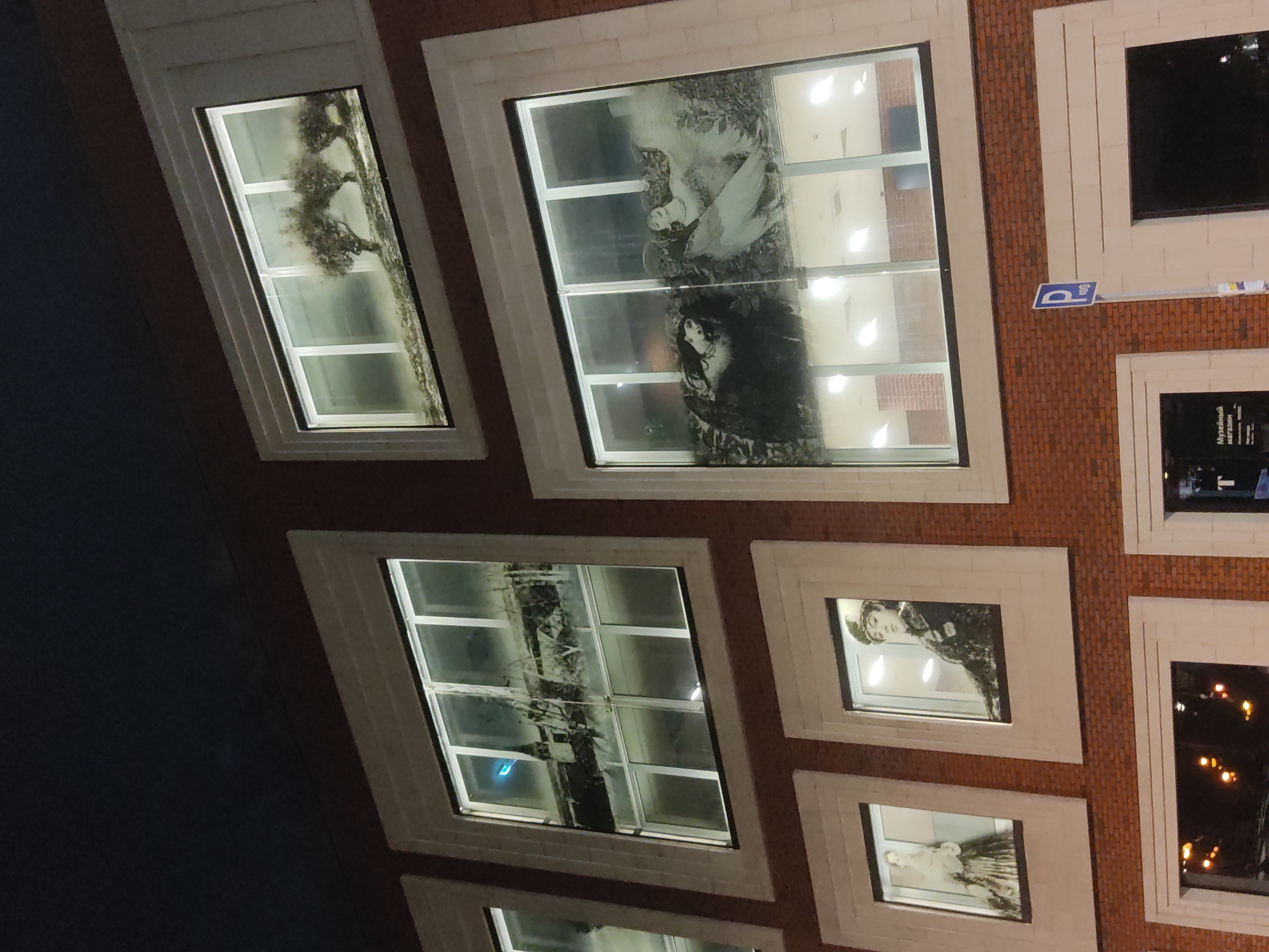 A low-angle, rotated photograph depicts a building facade with multiple windows in Moscow, Russia. The facade is constructed with orange brick and embellished with white, rectangular frames surrounding each window. The windows contain various images, including trees, portraits of people, and abstract shapes. The building's interior is visible through the windows as bright, white lighting. In the top right of the image, a blue and white parking sign is visible. The sky is dark, suggesting it is night. The photograph is rotated counter-clockwise.