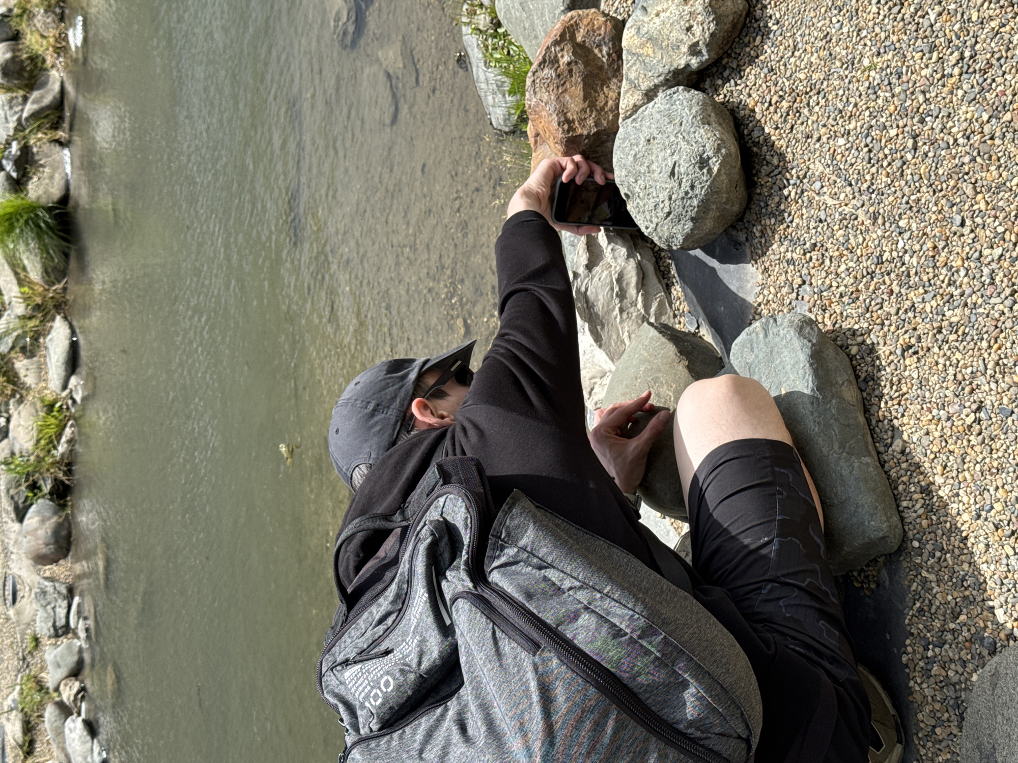 A person is photographed from an overhead angle, seated at the edge of a small river in a park or outdoor area. The person is wearing a black long-sleeved shirt, black shorts, a gray cap, and sunglasses. They have a gray backpack. The person is holding a smartphone in their extended right hand, seemingly taking a picture of the river or something across it. The river's edge is lined with rocks, and the ground around the person is covered with small pebbles. The location is likely in Estosadok, Russia.