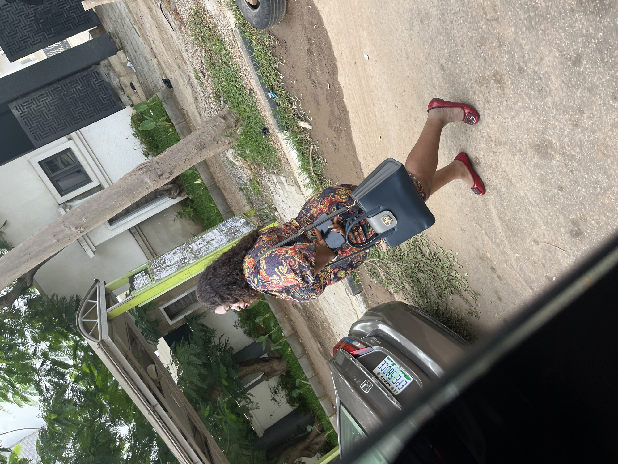 In Aso, Nigeria, a person wearing a colorful, patterned dress and red flat shoes walks alongside a grey car. The person carries a dark blue handbag. The scene is outdoors, with a paved surface and a grassy area. Green trees and foliage are visible, along with elements of building construction. A weathered wooden beam is in the foreground, and a tire is visible in the background. The license plate on the car reads "EPE 580CE LAGOS." The lighting suggests it is daytime.
