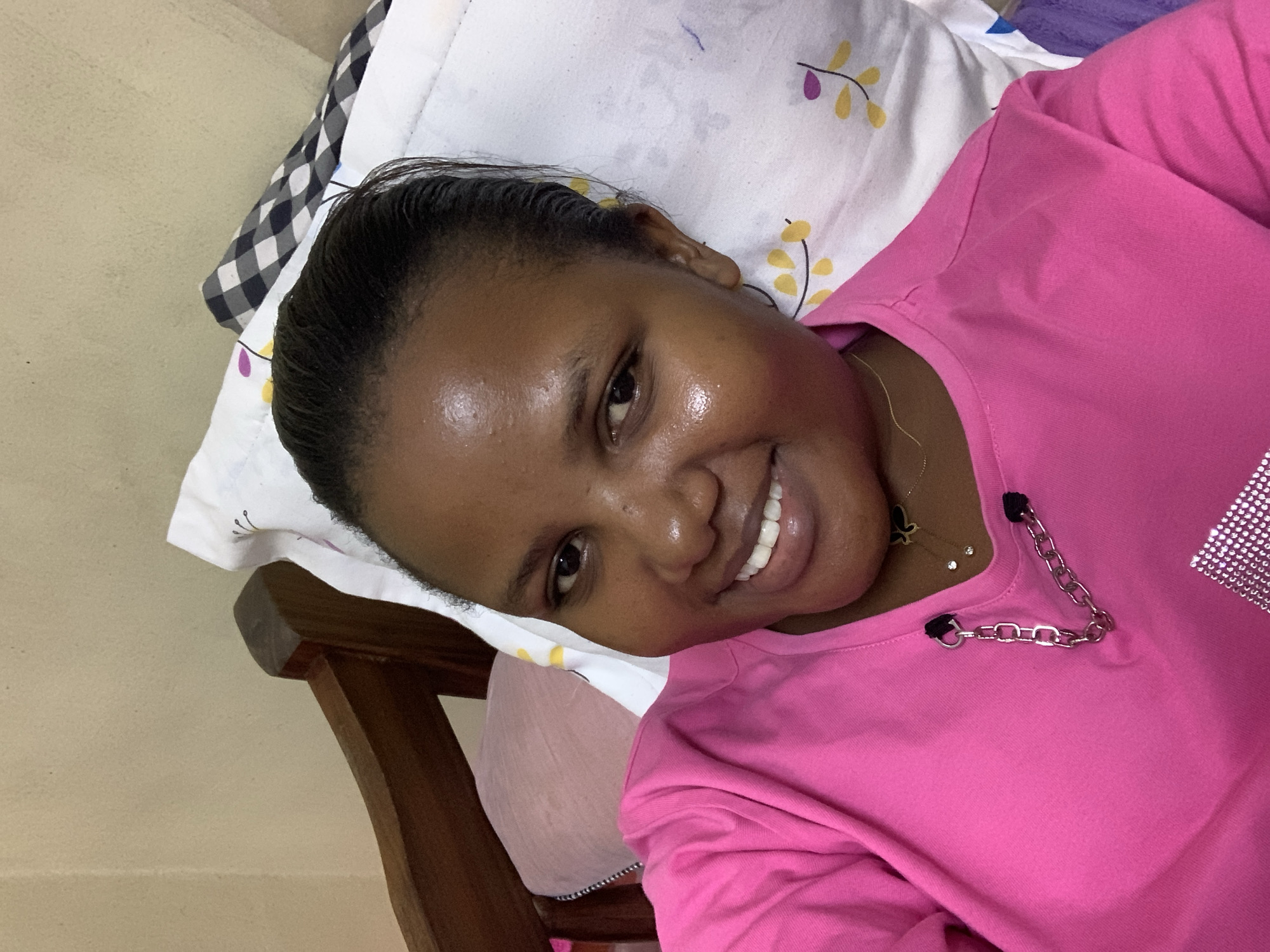 A young woman with dark hair pulled back and a pink shirt is lying on a pillow with a patterned cover. The woman is smiling at the camera. She has dark skin and a silver chain attached to her shirt collar. She also wears a gold necklace. The background shows a wooden chair and a beige wall. The image does not provide information about the location in Dar es-Salaam, Tanzania.