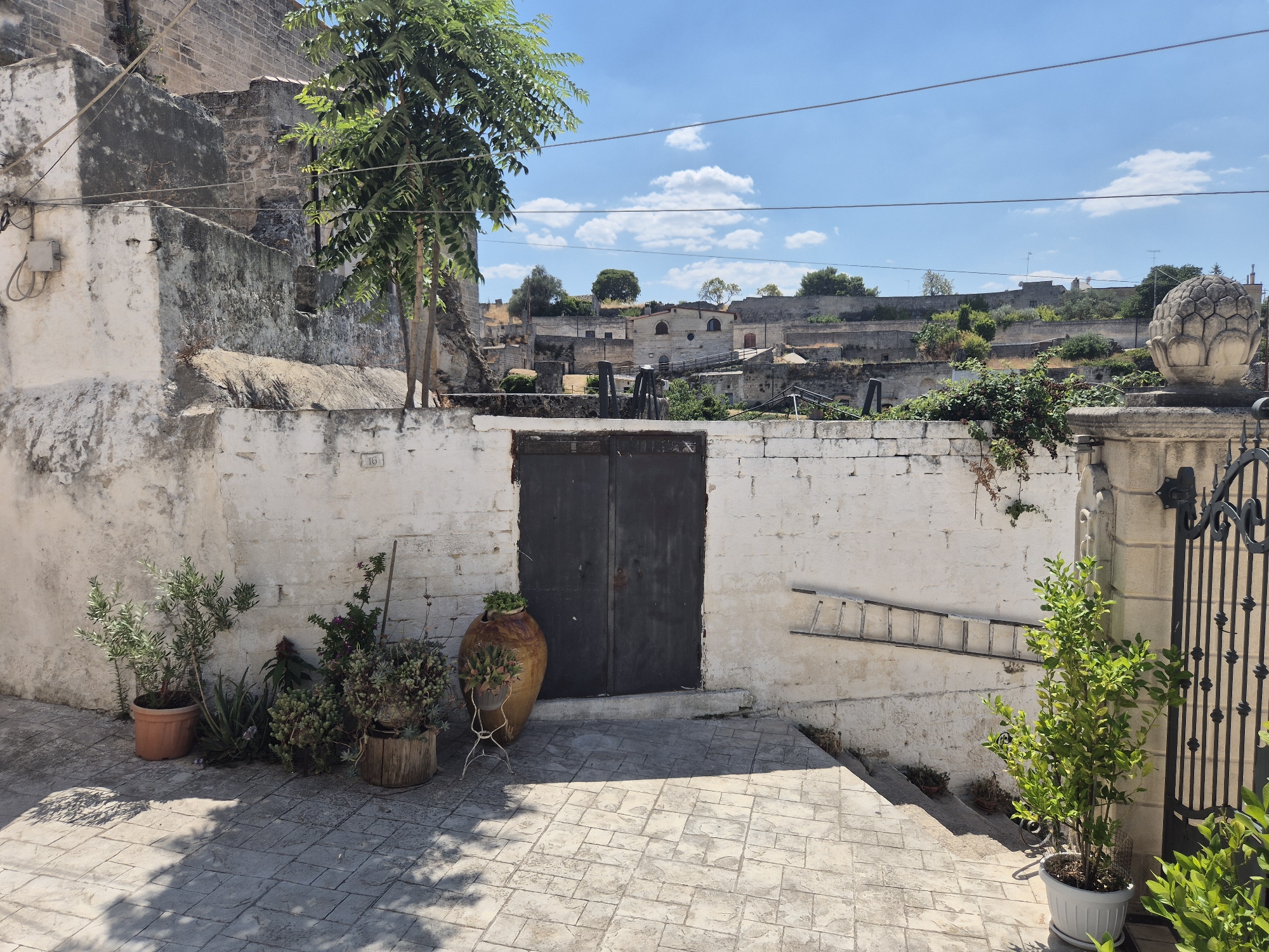 The image captures a street scene in Laterza, Italy, with a view of stone buildings and a blue sky with scattered clouds. In the foreground, a paved courtyard is lined with potted plants and a large ceramic vase. A dark double door is set into a white-washed stone wall. To the right of the door, a wooden ladder leans against the wall. In the background, more stone structures and greenery are visible, with a decorative stone finial on a pillar to the far right.