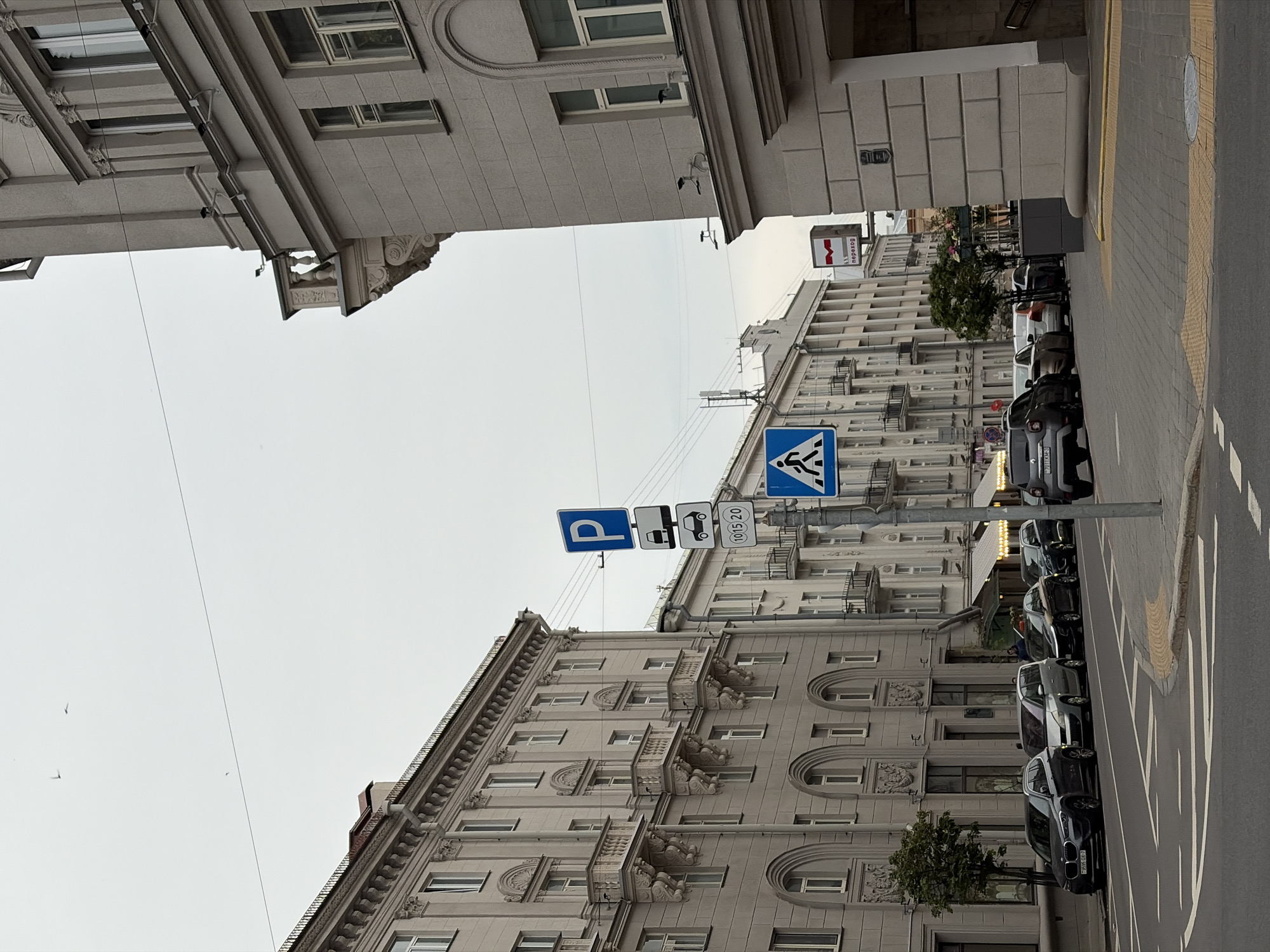 The image depicts a street scene on a cloudy day in Minsk, Belarus, specifically on Independence Avenue (Prospekt Nezavisimosti) near Oktyabrskaya Square (Kastrychnitskaya Ploshcha), looking eastward.
In the foreground, an asphalt road with white painted markings for traffic and a pedestrian crossing area with yellow tactile paving is visible. To the right, a grey paved sidewalk leads towards a large, light-grey building with a classical architectural style, featuring arched windows on the ground floor and an entrance recessed under an archway. A red "M" metro sign with the word "переход" (perekhod, meaning "underpass" or "transfer") in white text indicates a metro station entrance adjacent to this building. Security cameras are mounted on the facade.
On the left side of the street, a row of parked cars lines the curb. From left to right, a dark grey BMW X1 (Belarusian license plate 7858 OX-7), a silver Renault Espace, a dark sedan, another dark sedan, and a lighter-colored vehicle are visible. Behind these cars, another grand, multi-story building in a similar classical/Stalinist Empire style dominates the left side of the frame. This building features multiple ornate balconies with sculpted supports and decorative cornices. A small tree with green foliage is planted on the sidewalk in front of this building. Further down the street, on the right side, another dark grey Renault Duster (Belarusian license plate 7011 XM-3) is parked.
In the center of the image, a light-colored pole supports several blue and white road signs. The topmost sign is a blue square with a white "P" for parking. Below it are two white rectangular signs: one shows a car and a small truck, likely indicating allowed vehicle types, and the other shows a car with "10-15-20", possibly a parking tariff or time limit. Below these, a blue triangular sign with a white walking figure indicates a pedestrian crossing. Overhead wires traverse the upper portion of the scene. The sky is overcast and light grey, with no direct sunlight. Three small birds are visible high in the sky. Minimal human presence is discernible; a few indistinct figures can be seen in the distance between buildings and parked cars.