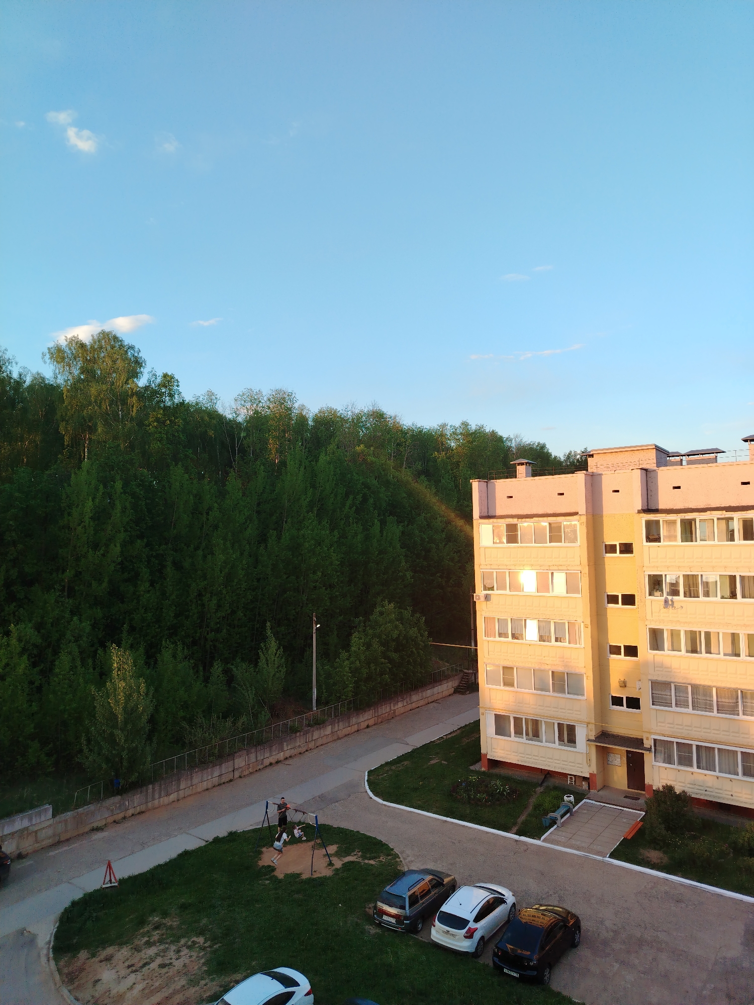 The image shows a residential scene in Noviy Urengoy, Russia. A multi-story yellow and pink building is visible on the right, with numerous windows reflecting sunlight. To the left of the building is a dense forest. In the foreground, there's a paved area with several parked cars: a blue compact car, a white sedan, and a black sedan. A small playground area with swings is present, where adults and children are interacting. A concrete wall separates the playground from the road. The sky is blue with scattered clouds.