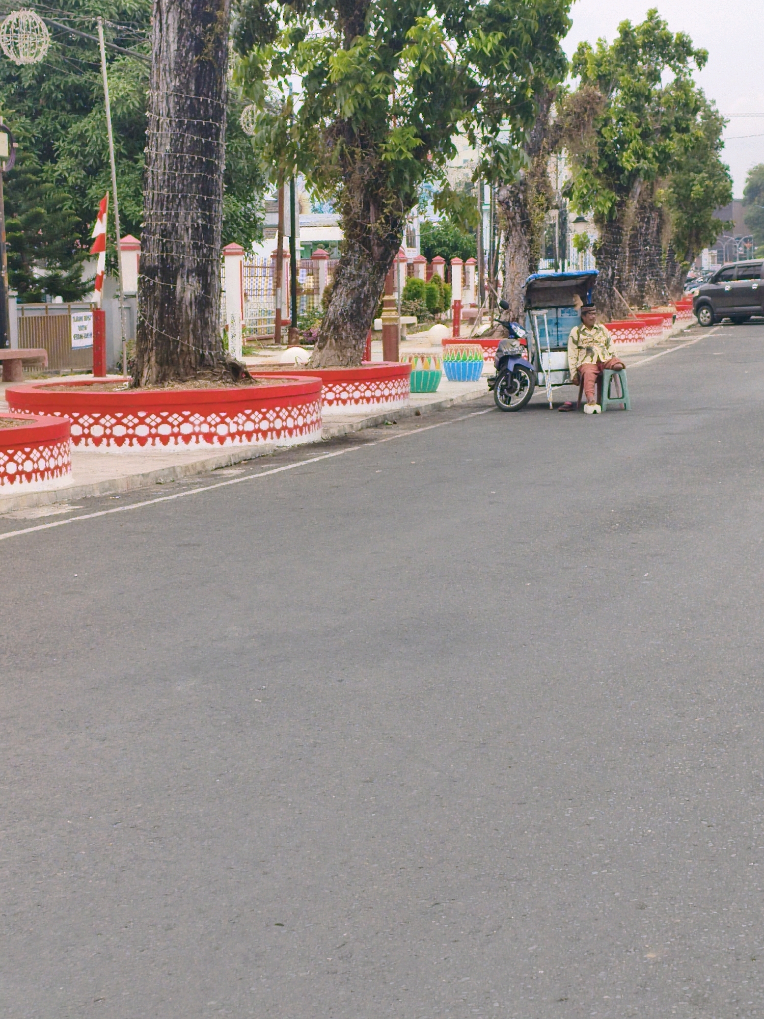 A street scene in Bengkulu, Indonesia, features a man sitting on a green plastic stool next to a three-wheeled motorcycle-taxi, commonly known as an "ojek" or "becak motor." The man is wearing a light-colored patterned shirt and reddish-brown pants, with his feet on the ground. He appears to be waiting, possibly for a passenger. The ojek is parked on the asphalt road, with a blue canopy over its passenger area. In the background, a row of trees lines the sidewalk, which is adorned with red planters decorated with white geometric patterns. Behind the trees, a fence with colorful elements and a building can be seen. A dark-colored SUV is visible on the far right of the road. The sky is overcast.