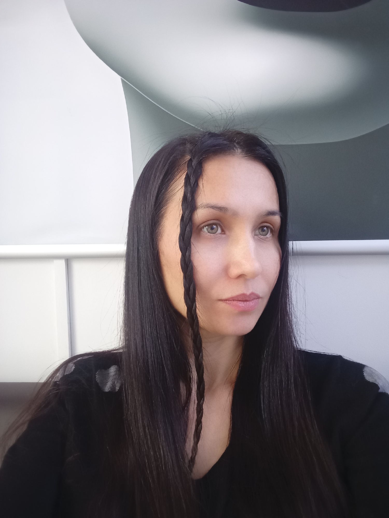This is a medium close-up portrait of a woman photographed indoors. She has long, dark, straight hair, featuring two distinct braids that frame the left side of her face. Her light-colored eyes, appearing hazel or light green, are cast slightly upwards and to her right, suggesting a contemplative or observant gaze. Her expression is calm and composed, with natural, light pink lips. She is dressed in a dark, long-sleeved top, likely black, which exhibits subtle lighter, possibly patterned or reflective, detailing on the right shoulder.
The background is minimalist and modern, characterized by a smooth, curving surface in shades of white and light grey, with darker grey sections towards the top. A straight white bar or ledge extends horizontally across the frame behind her. The lighting is soft and diffused, typical of an indoor setting. No specific visual cues within the image confirm the provided location context of Izhevsk, Russia. There is no visible text, and the time of day or external weather conditions cannot be determined from the photograph.