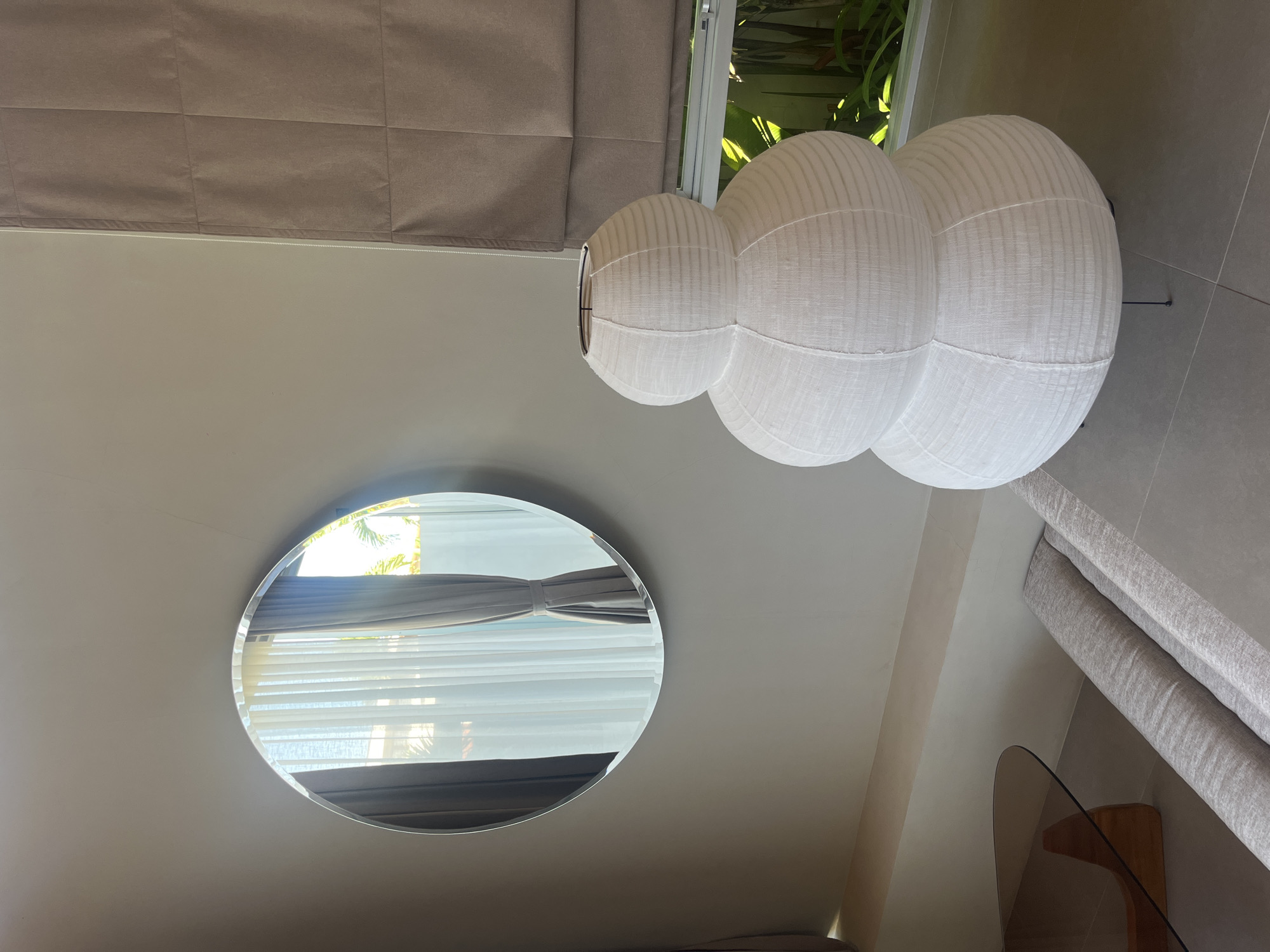 The photograph, taken indoors, presents a partial view of an interior space, likely within a building located in Kuta Selatan, Indonesia. The scene is composed of a gray tiled floor. On the top left of the frame there is a light brown curtain hanging down. A white paper lantern sculpture is shown, composed of stacked bulbous segments. A circular mirror on the wall reflects a window with light brown blinds and a glimpse of green foliage outside. The furniture is gray and tan. Overall, the image focuses on interior design elements with a neutral color palette.