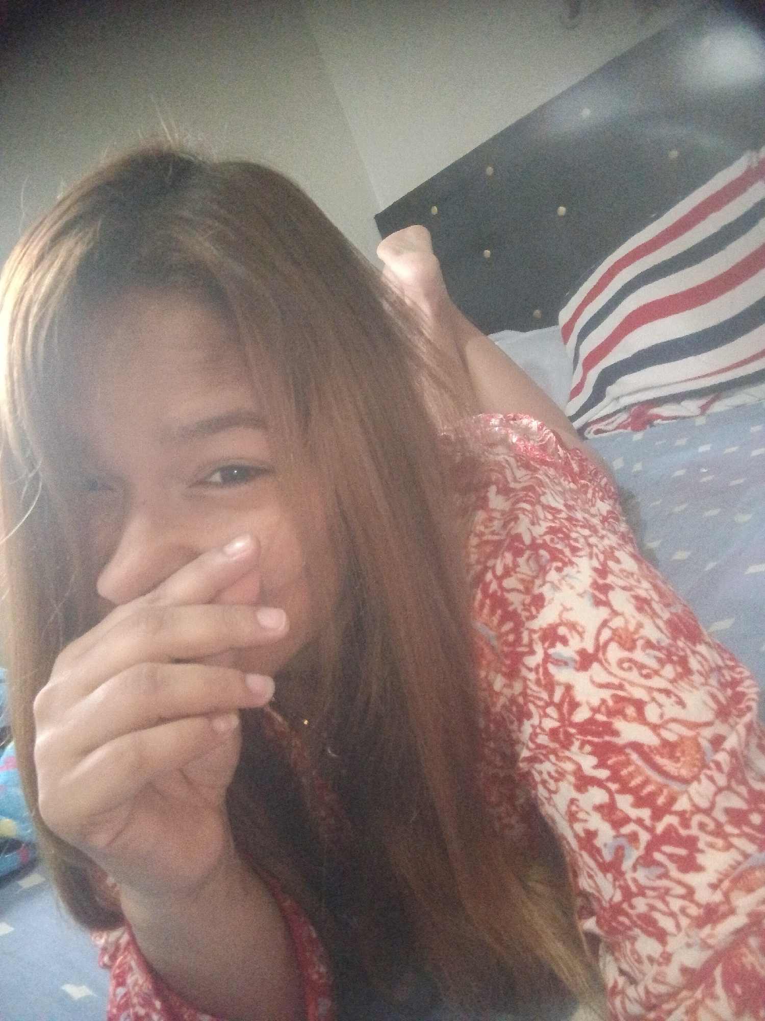 A person with long, brown hair is lying on their stomach on a bed. They are wearing a red and white patterned top and covering their mouth with their hand. Their head is angled towards the camera. A striped pillow is visible behind them against a dark headboard. The bed has a light blue patterned sheet. There is no information in the image to indicate that this image was taken in the city of Caracas, Venezuela.