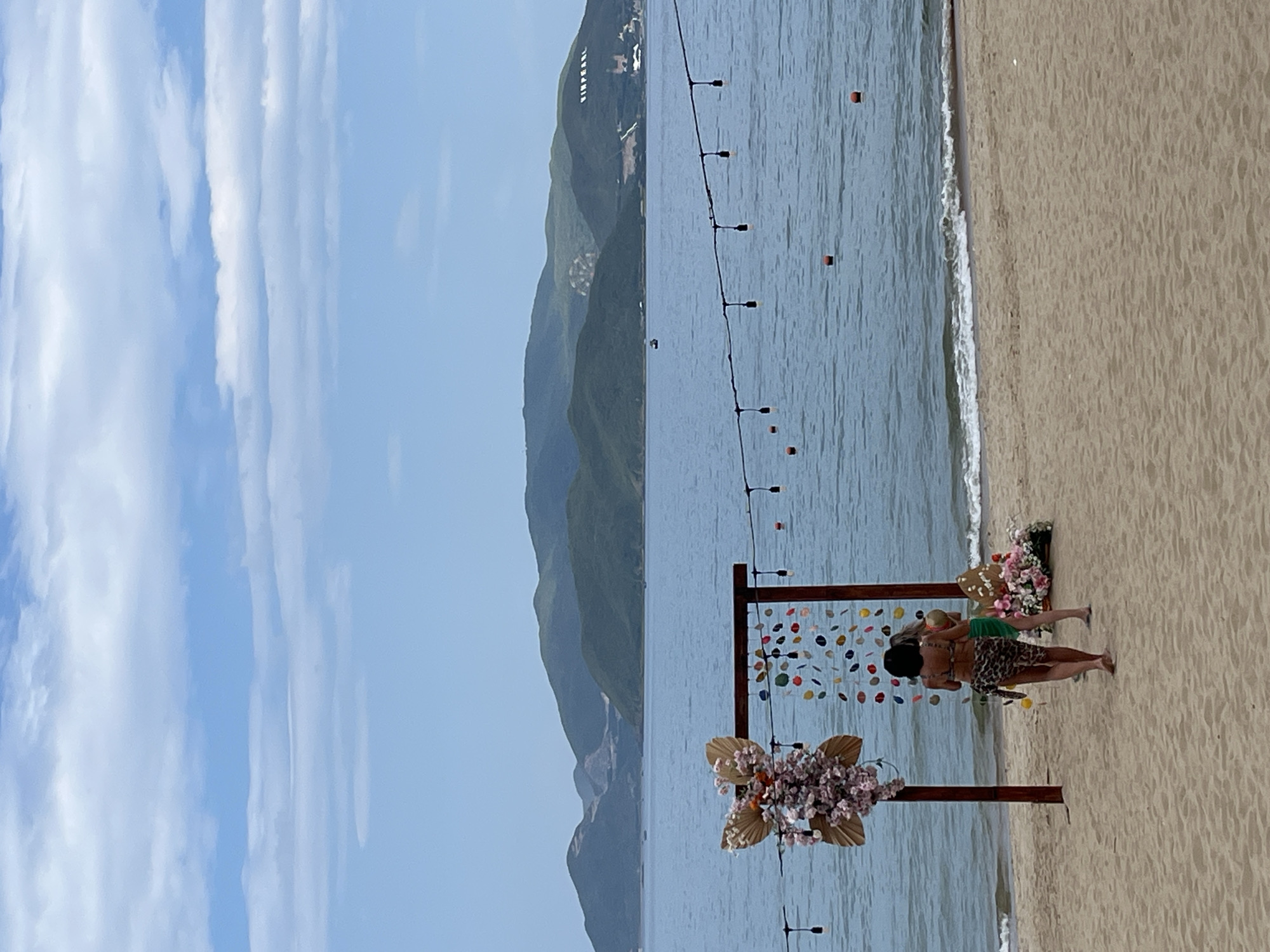 Two women are on a sandy beach in Nha Trang, Vietnam, near a decorative archway adorned with flowers and seashells. The archway is positioned in front of the calm, blue ocean, with lush green mountains in the background. A string of lights is suspended above the water, and several buoys are visible on the surface. The woman in the foreground wears a leopard-print bikini with a matching sarong, while the woman behind her is in a green bikini. The overall scene suggests a relaxed, tropical atmosphere.