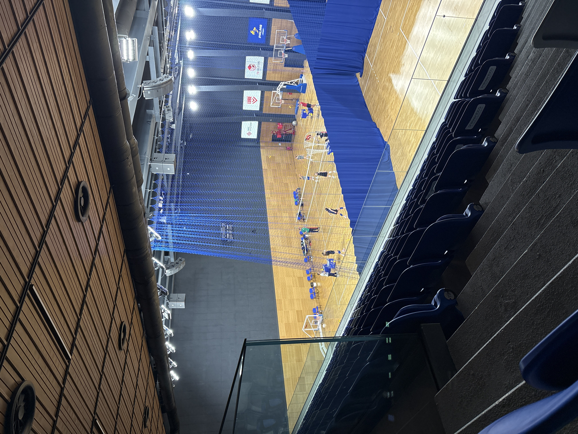 The image shows an indoor volleyball court located in Sofia, Bulgaria. The court is made of polished wood and is surrounded by blue netting. Several people are playing volleyball, including two individuals on the court with a net and others observing. The stands are filled with empty blue seats, indicating it is not an active event. The ceiling is made of wood panels and industrial piping, with lighting fixtures visible. The overall impression is that of a modern sports facility, likely used for training or local games.