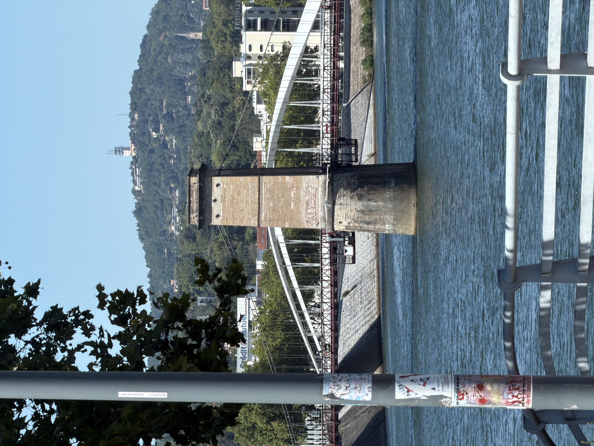 The image depicts a view of the Rhône River in Lyon, France, with a bridge and its supporting tower prominently featured. The perspective is from a low angle, looking upwards and slightly sideways, with the scene rotated.
In the foreground, a large, cylindrical, stone-built tower stands as a support for a suspension bridge. The tower is weathered and shows signs of graffiti. To the right, a metal railing with vertical bars and a curved support is visible, suggesting a walkway or part of the bridge structure.
The Rhône River flows to the right of the tower, its surface rippled and appearing a cool blue-green. The far bank of the river is lined with buildings, some appearing modern with large windows, and others older in architectural style. Behind the buildings, a densely forested hill rises, dotted with what seem to be scattered structures and a visible communication tower or structure near the summit.
The bridge itself is a suspension bridge with a large white curved arch and a latticework structure of red girders and white supports. The presence of trees with green leaves suggests it is a warm season, likely summer or late spring. The sky above is a clear, bright blue, indicating a sunny day.
Visible text on the main image includes graffiti on the tower, some stickers on a pipe in the foreground, and the partial word "Crédit mutuel" on a building in the background, a French mutual banking group. The overall impression is a sunny day in a European city, showcasing an urban landscape alongside natural elements.