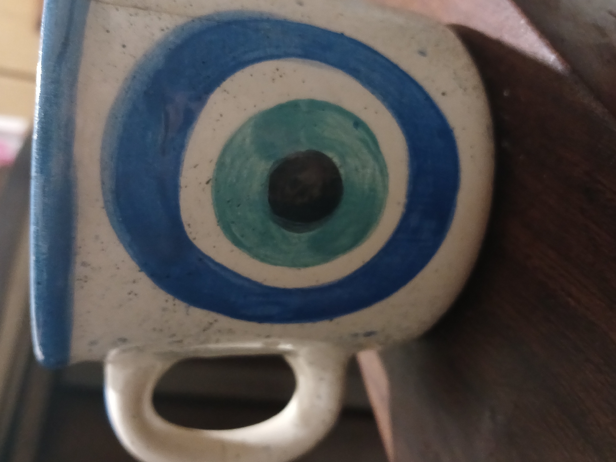 The image shows a close-up of a white ceramic mug decorated with concentric circles in blue and turquoise, resembling an eye. The mug has a handle on the bottom left side. The mug is placed on a dark brown wooden surface. The location is undetermined.