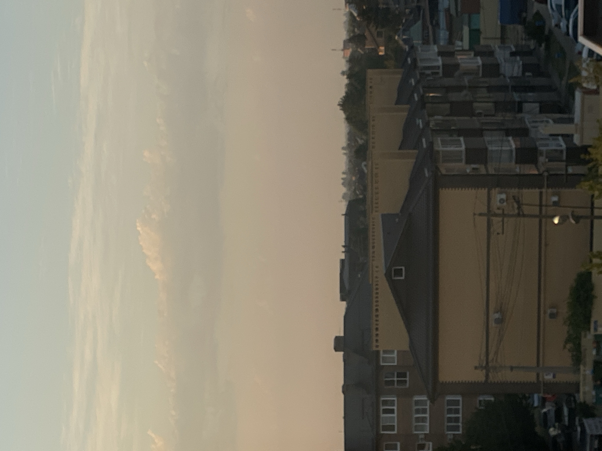 The image shows a cityscape with residential buildings under a hazy, pale orange sky. The buildings are multi-story structures, some with dark gray roofs and others with tan facades. Windows are visible on the buildings, some with white frames. There are visible power lines and what appear to be air conditioning units attached to the sides of some buildings. In the foreground, there are trees and parked cars. The background is filled with more buildings and trees, suggesting a densely populated area. The overall impression is of an urban environment, possibly during dawn or dusk, with diffused light.