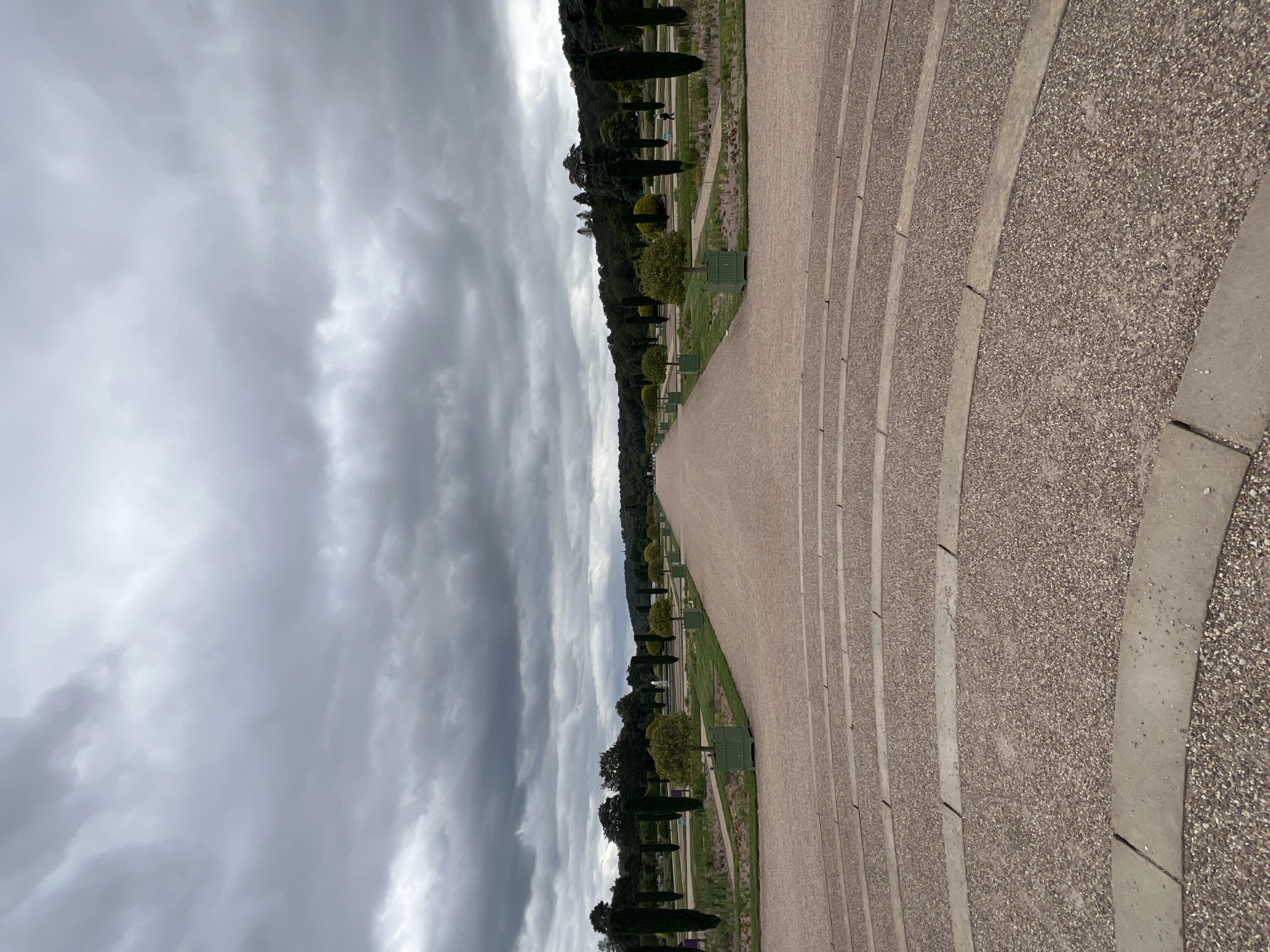 The image depicts a long, wide gravel pathway leading into a verdant garden landscape. The pathway is flanked by manicured trees and shrubs, some of which are contained in green square planters. In the distance, a fountain is visible, and beyond that, a line of dense green trees forms the horizon. The sky above is overcast with heavy grey clouds. In the foreground, there are curved, tiered sections of gravel, appearing to be part of a grand entrance or viewing area. The overall impression is of a formal, expansive garden setting. The location is not Stafford, United Kingdom; it is the Gardens of Versailles, France.