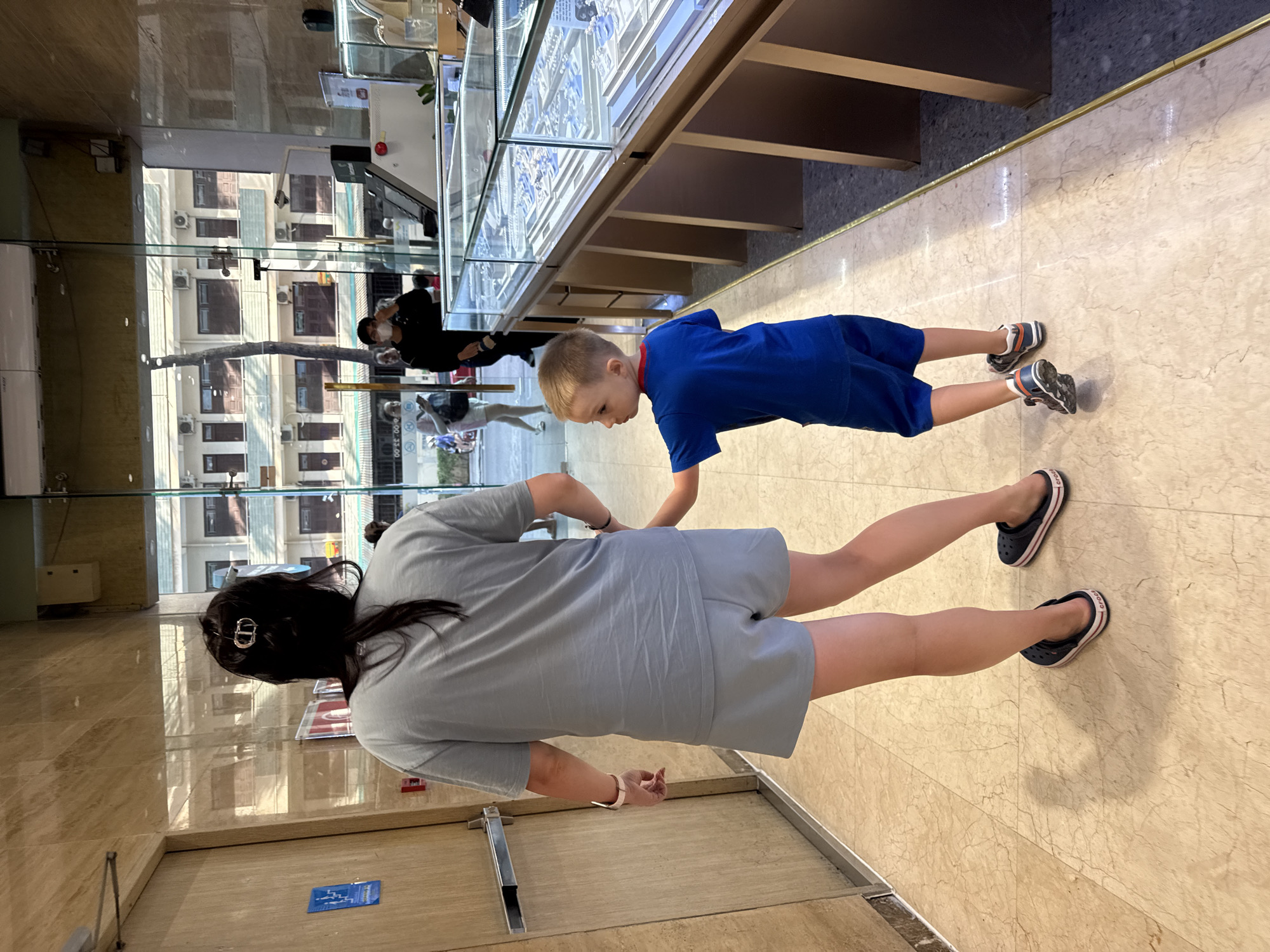 A woman and a young boy are walking in a jewelry store in Nha Trang Ward, Vietnam. The woman is wearing a gray t-shirt and shorts, and the boy is wearing a blue t-shirt and shorts. They are holding hands as they walk past display cases filled with jewelry. The store appears to be modern and well-lit, with marble floors and glass display cases. Outside the store, a street scene is visible through the glass, with buildings and trees.