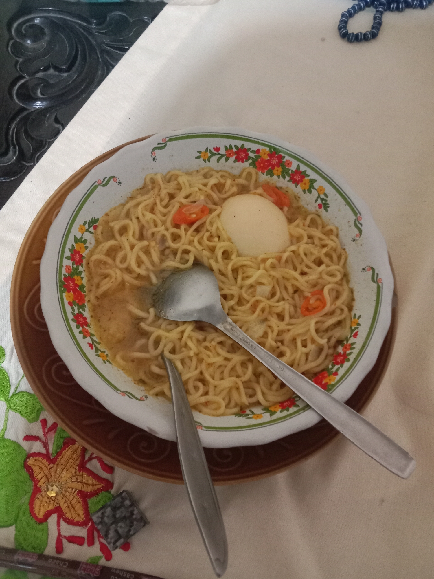 A close-up, overhead shot shows a bowl of instant noodles with a hard-boiled egg and sliced chili peppers. The noodles are golden yellow and appear to be in a broth. A spoon rests on top of the noodles, and a fork is positioned to the right of the bowl. The bowl is white with a floral rim in red, yellow, and green. It sits on a brown saucer, which is placed on a beige tablecloth with embroidered floral patterns in green, red, and yellow. In the upper right corner, a strand of dark blue beads is visible. In the lower right corner, a wrapper for a chocolate bar with the word "Choco" and "Cashew" printed on it is partially visible. To the left of the bowl, a dark, carved decorative wooden piece is present, and behind it, a turquoise object is visible. The scene appears to be a casual meal setting. The location is likely a residence in Madiun, Indonesia, given the cultural context of instant noodles being a common meal and the presence of local food packaging.