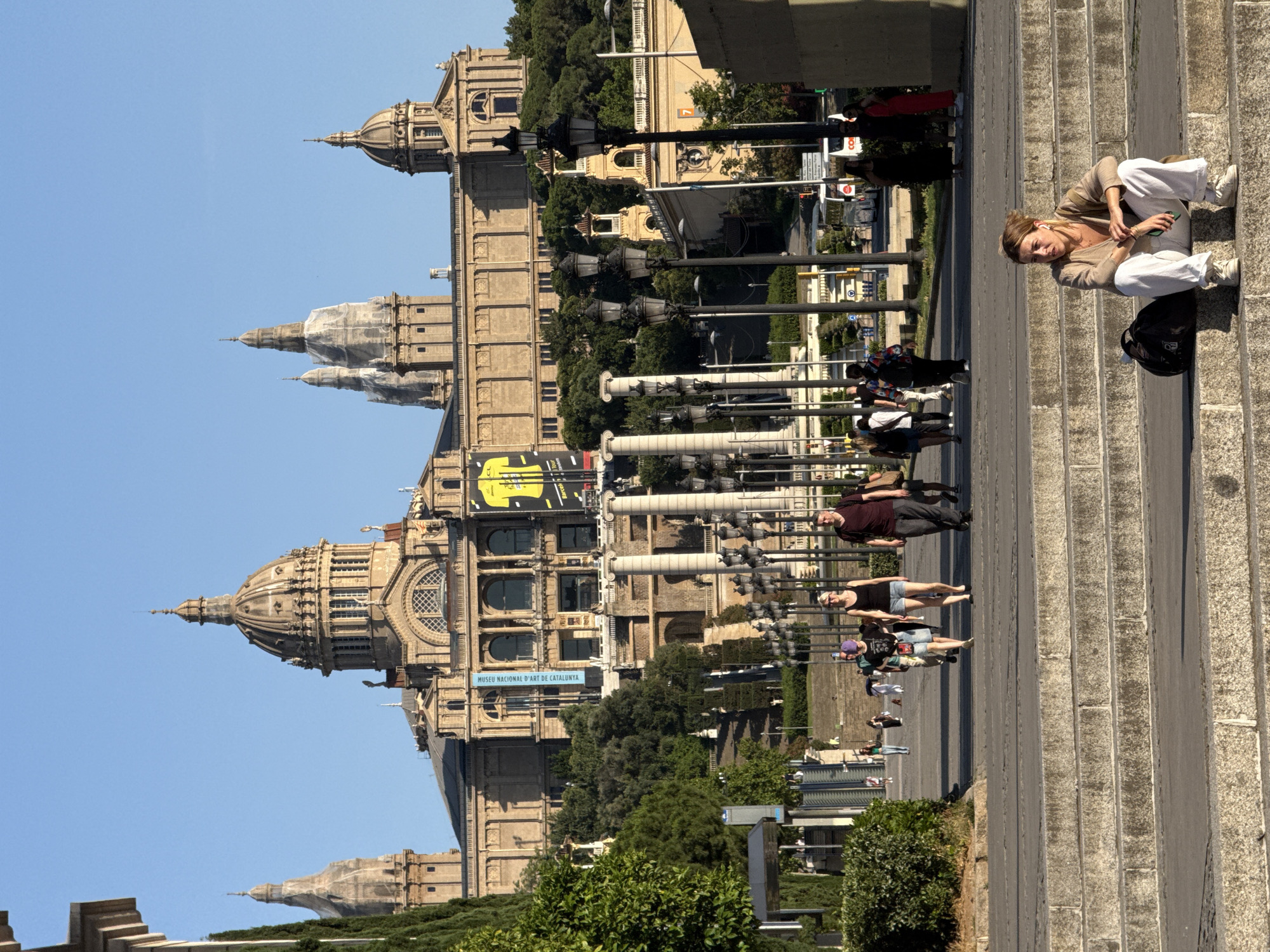The image depicts a view of the Palau Nacional, the National Palace of Catalonia, situated on Montjuïc hill in Barcelona, Spain. The grand building, characterized by its prominent domes and classical architecture, dominates the background under a clear blue sky. In the foreground, a wide stone staircase leads up towards the palace, with a woman seated on the steps, looking off to her right. She is wearing a beige cardigan, white pants, and white sneakers, with a black backpack beside her. Several people are walking on a paved pathway in front of the stairs, approaching the palace. The pathway is lined with lampposts and trees. The overall scene captures a sunny day with tourists and locals enjoying the view of the iconic Barcelona landmark.