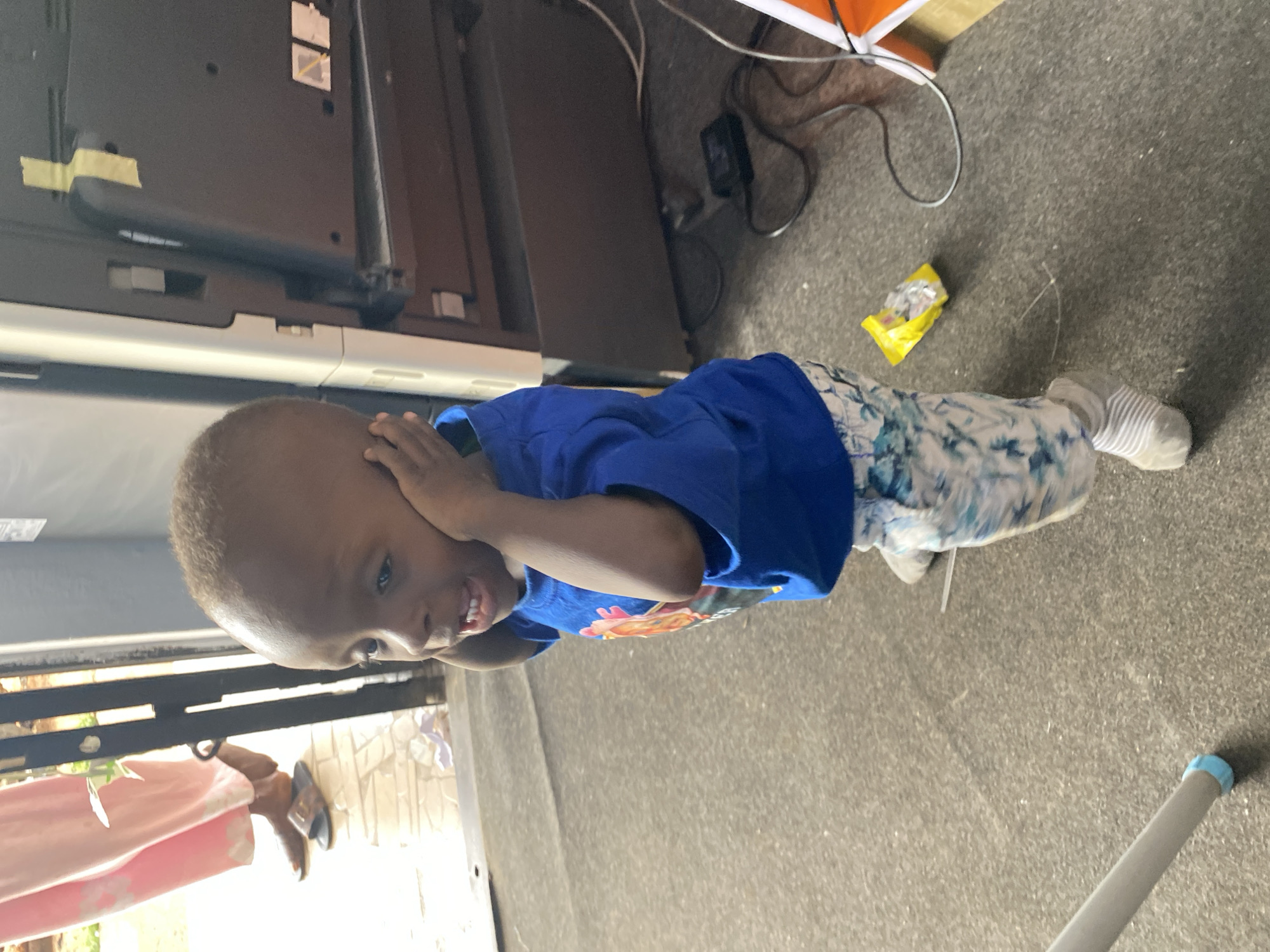The image features a young child, likely a toddler, standing indoors. The child has dark skin and short, light-colored hair, and is wearing a royal blue t-shirt with a colorful graphic on the chest and light-colored patterned trousers. White socks with grey stripes are visible on their feet. The child is captured mid-action, with both hands pressed against their ears or sides of the head, and a wide, joyful smile revealing their teeth.
The scene appears to be an indoor space with a grey, textured floor, possibly carpet or concrete. In the background to the left, a large, dark flat-screen television or monitor is visible, possibly mounted, with some yellow tape on its back. Electrical cables are scattered on the floor around this equipment. To the child's right, a crumpled yellow wrapper, resembling a snack or candy wrapper, lies on the floor next to more cables. A light-colored leg of a piece of furniture or a stand with a light blue cap is partially visible in the bottom right corner.
Beyond the child, through what appears to be a doorway or opening, another person's leg is visible, dressed in a light pink garment and brown sandals. Some light-colored, patterned brickwork or a wall is also discernible in this area. The bright lighting suggests it is daytime. The child's pose and expression convey a sense of playfulness or amusement. No discernible text is visible on any objects in the image.