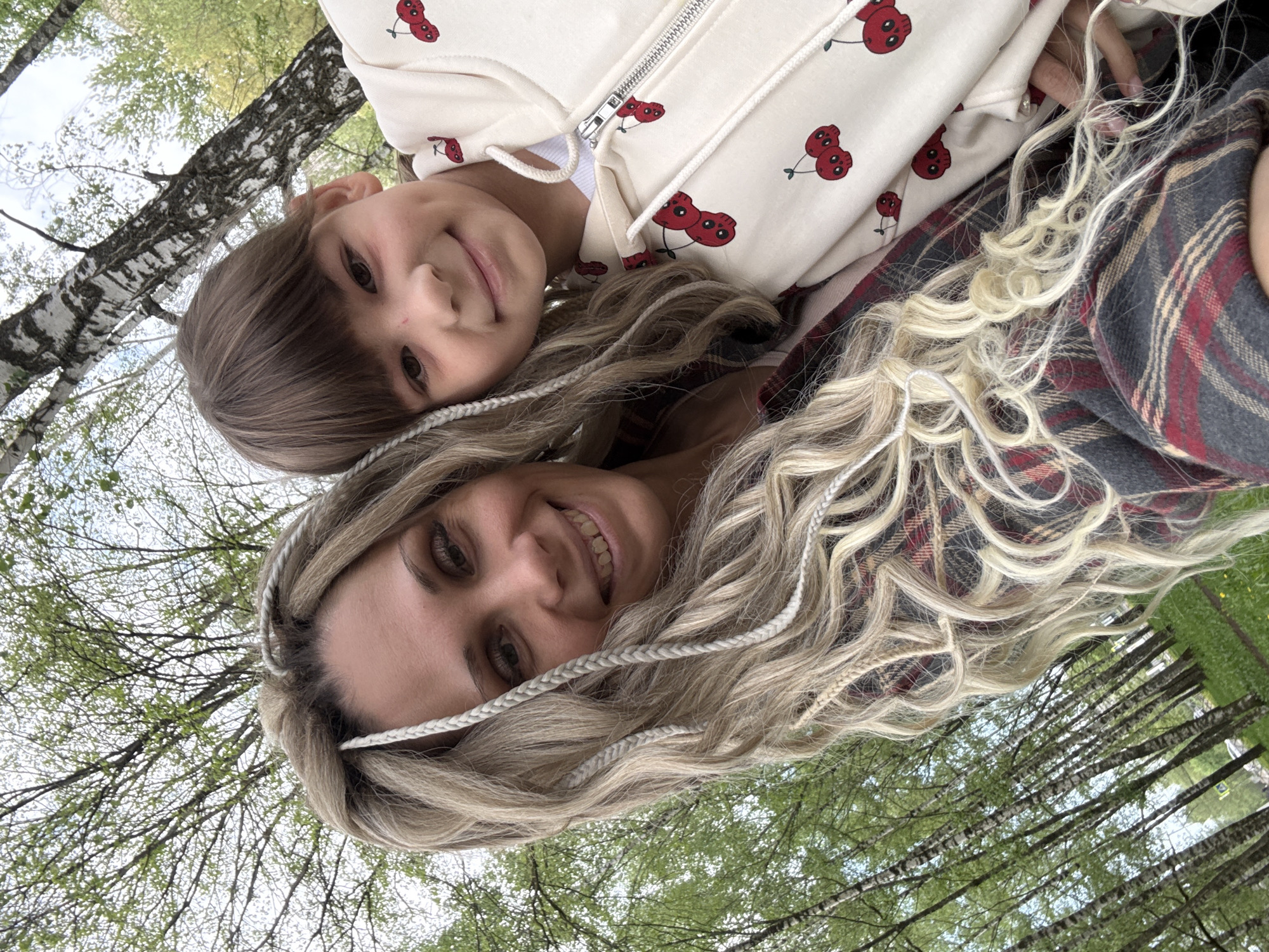The image depicts a selfie taken outdoors, likely in a park, featuring a woman and a child. The woman has blonde hair styled with decorative braids, and is wearing a plaid garment. The child has brown hair and a cherry-patterned jacket. The background shows trees, suggesting a natural setting. The photo was most likely taken in Rusanovo, Russia.