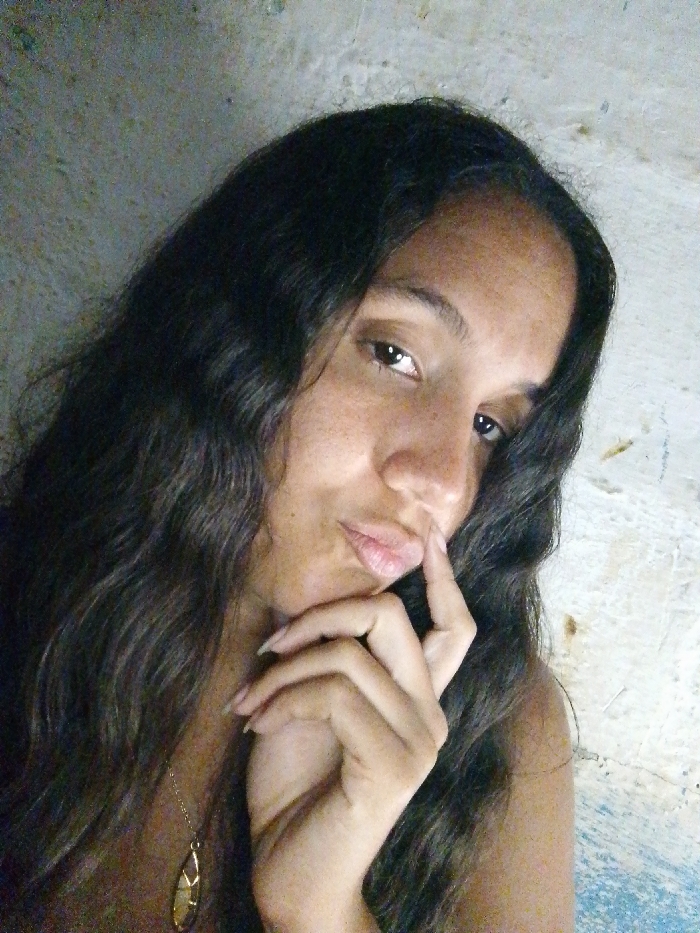 A portrait-style image depicts a young female indoors, presumed to be in Juan Griego, Venezuela. The subject has dark, wavy hair extending past her shoulders, dark eyes, and a medium complexion with visible freckles across her face. She is looking directly at the camera with a "duck face" expression, her lips slightly pursed. Her right hand is raised, with index and middle fingers touching the area just below her lower lip. She is wearing a delicate gold-toned necklace with an oblong, abstract pendant visible on her chest. Her left shoulder is bare, indicating a sleeveless garment or a wide neckline. The background consists of a textured, light-colored wall with visible variations in tone and surface irregularities, consistent with painted or unpainted concrete or stucco. The lighting is moderate, casting soft shadows.
