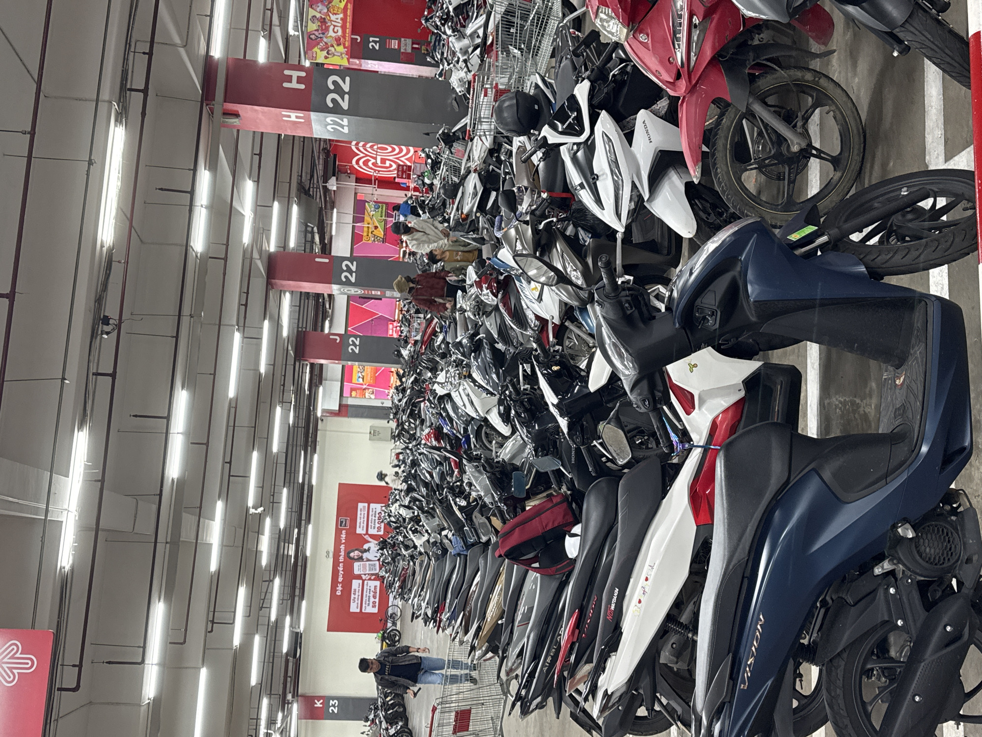 The image depicts a crowded indoor motorcycle parking area, likely a basement or covered lot, filled with numerous scooters and motorcycles. The scene is brightly lit by fluorescent lights on the ceiling. Several pillars with large numbers and letters ("K 23", "H H 22", "J 22") are visible, suggesting a structured parking system. The ground is concrete, with white lines marking parking spaces.
In the foreground, a dark blue scooter with "VISION" written on its side is prominent. To its right, a red scooter is partially visible. Further back, a white Honda scooter is parked. There are several people in the image, mostly walking or standing near their vehicles. One man in a dark jacket and jeans is walking towards the left, holding a phone. Another group of three people, two adults and a child, are seen in the middle ground. The overall impression is one of a busy public parking facility.
While the image has a sign with Vietnamese text and a distinctly Southeast Asian aesthetic often associated with Da Lat, Vietnam, there are no explicit visual cues within the image that definitively pinpoint the location to Da Lat. Therefore, stating the location as Da Lat, Vietnam, is an assumption.