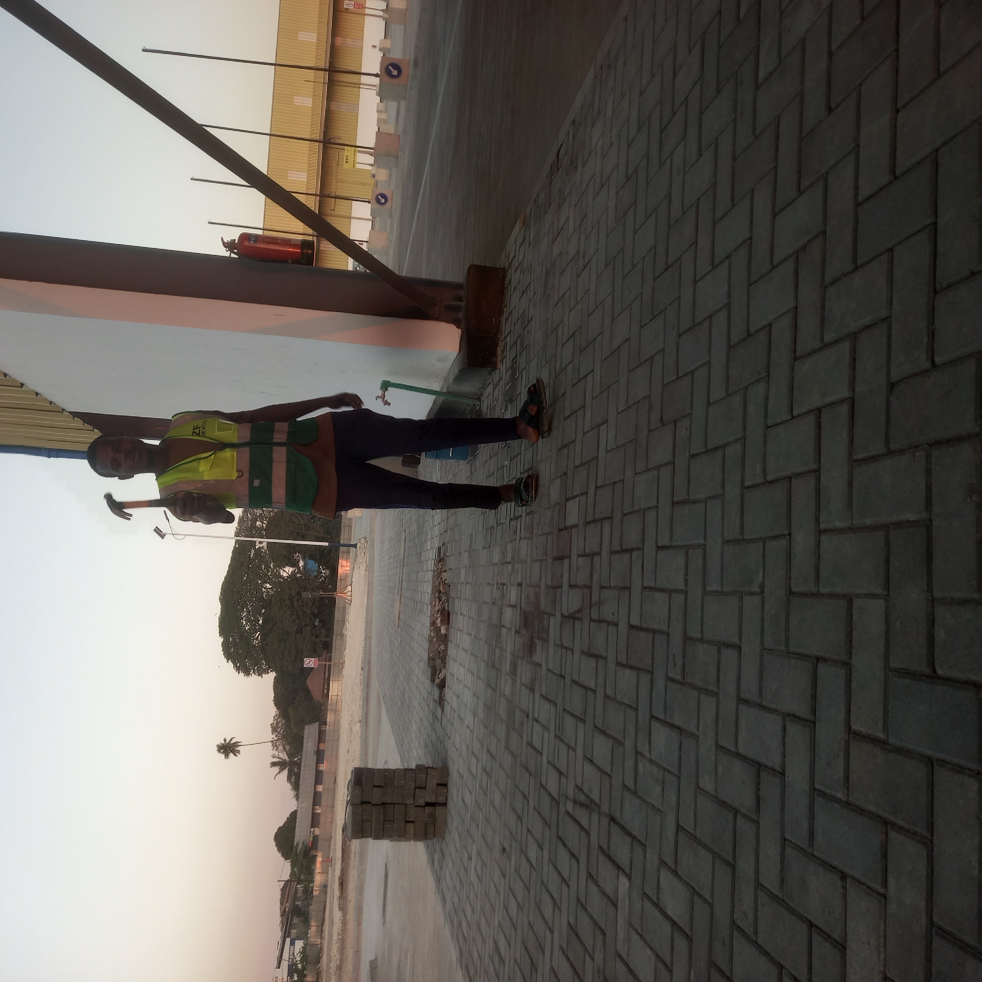 A medium shot captured at ground level shows a male construction worker holding a hammer in his right hand, extended towards the camera, in Bububu, Tanzania. He is wearing a green reflective vest over a brown shirt and dark pants, with black sandals. The worker is standing on a paved walkway composed of interlocking grey rectangular bricks laid in a herringbone pattern. Behind him, a stack of identical bricks is visible to his left. Further in the background, a large building with a yellow facade is partially visible, with a red fire extinguisher mounted on its exterior wall. Trees and a clear sky complete the scene.