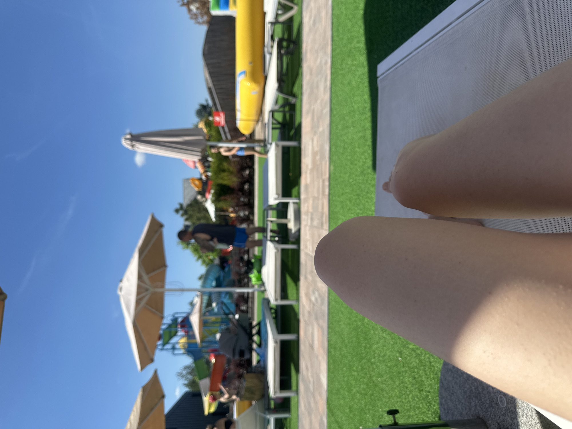 The image depicts a sunny day at an outdoor water park or amusement area in Khotianivka, Ukraine. The perspective is from someone reclining on a lounge chair, focusing on their legs which occupy the foreground. In the background, a bright blue sky suggests fair weather.
Several objects are visible, including lounge chairs arranged on a paved area next to a green, artificial turf surface. A large, yellow inflatable slide is prominent, with other colorful attractions of the park, such as slides and structures, partially visible further in the background. Umbrellas, likely for shade, are also scattered around the area.
Several people are present, though most are out of focus. One person, wearing blue shorts, is seen standing near the yellow slide. Other figures are indistinct, suggesting activity and movement within the park. The overall scene conveys a sense of leisure and recreation on a warm, clear day. The angle of the sun suggests it is likely mid-morning to early afternoon. No visible text is discernible in the image.
