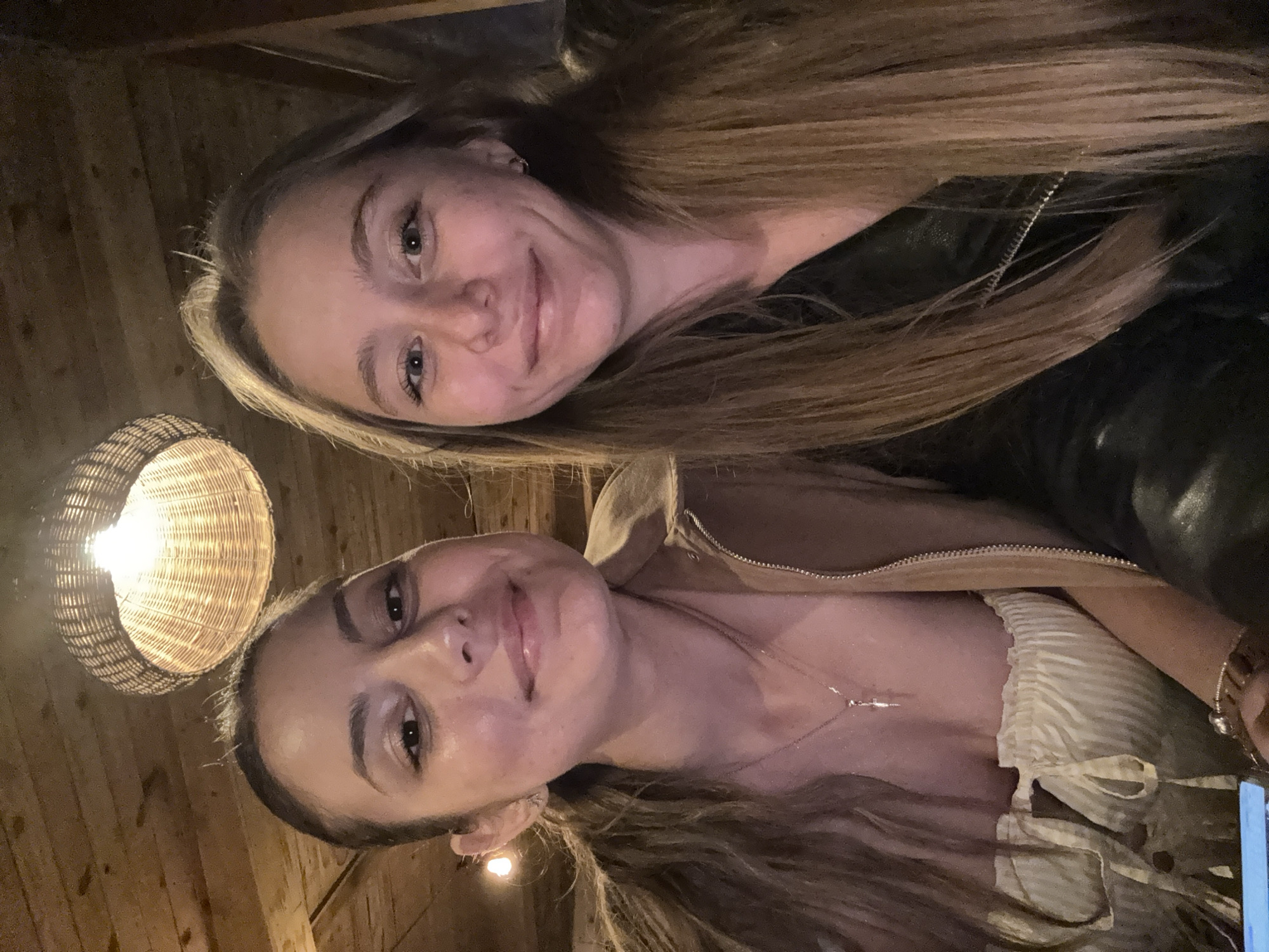 Two young women are taking a selfie in what appears to be a dimly lit restaurant with wooden paneling on the ceiling. A woven lampshade with a light bulb is visible in the upper center of the image. The woman on the left has her hair pulled back in a ponytail and is wearing a light-colored, strapless top with a necklace. The woman on the right has long, blonde hair and is wearing a black jacket. Both women are smiling at the camera. There are no other people or objects clearly visible in the image that would indicate a specific location in Tsarevo, Bulgaria.