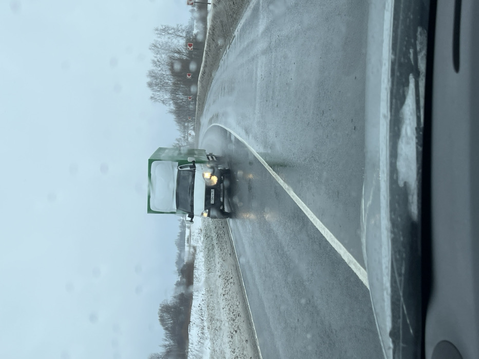 A white GAZelle-type light truck with a high, green box body is observed driving on a wet, asphalt road, seen from the perspective of a vehicle's dashboard and windshield. The truck's headlights are illuminated, and its Russian license plate, "Х584КХ 98," is visible. The road surface is visibly wet from precipitation, with a solid white line delineating the left side of the lane. The road curves sharply to the right in the mid-ground, indicated by red and white chevron signs positioned on the right shoulder. Snow-covered verges line both sides of the road, and the background features sparse, leafless trees and shrubs under an overcast, light grey sky. This scene occurs on a road in Rogachovo, Russia, during cold and damp weather conditions.