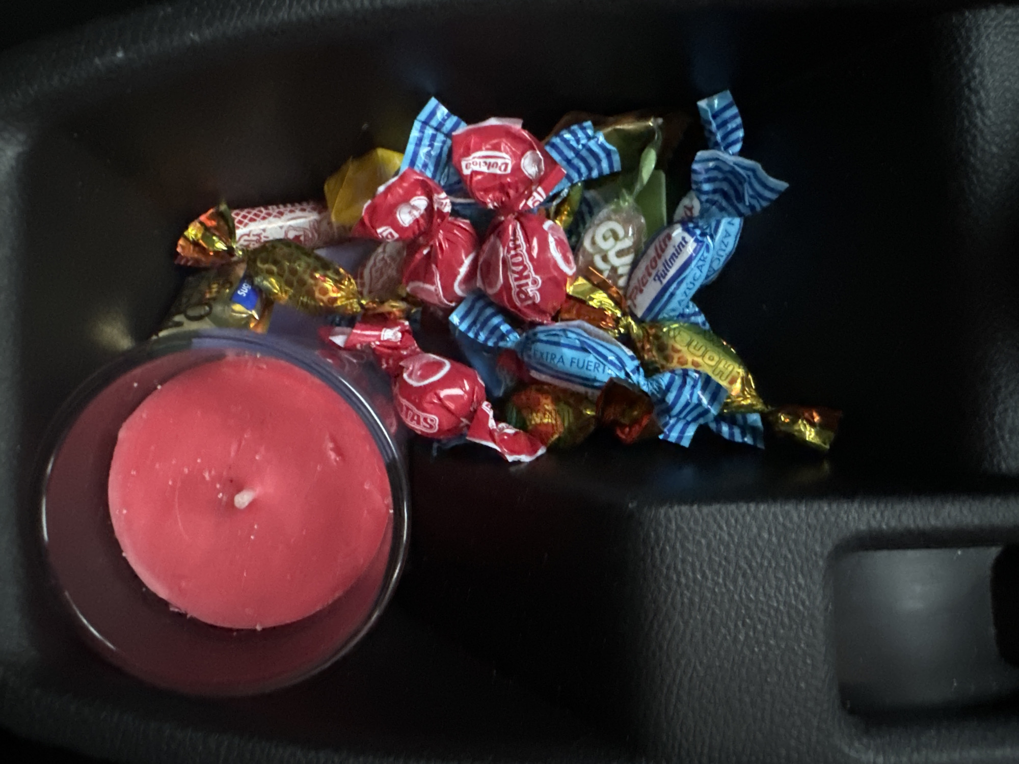 The image shows a close-up, overhead view of a red candle in a glass jar, placed within a dark-colored car's interior, likely a center console storage area. The candle is partially burned, with a visible wick. Surrounding the candle are several individually wrapped candies with brightly colored wrappers, predominantly red, blue, and gold. Some of the wrappers have text, including "PIKOT", "Dukino", and "Fullmint", suggesting they are mint or fruit flavored sweets. The car's interior appears to be textured, dark plastic. The scene is static and there are no people or actions depicted. The lighting is dim, consistent with the interior of a vehicle. There is no specific information in the image to suggest a location in Murcia, Spain.