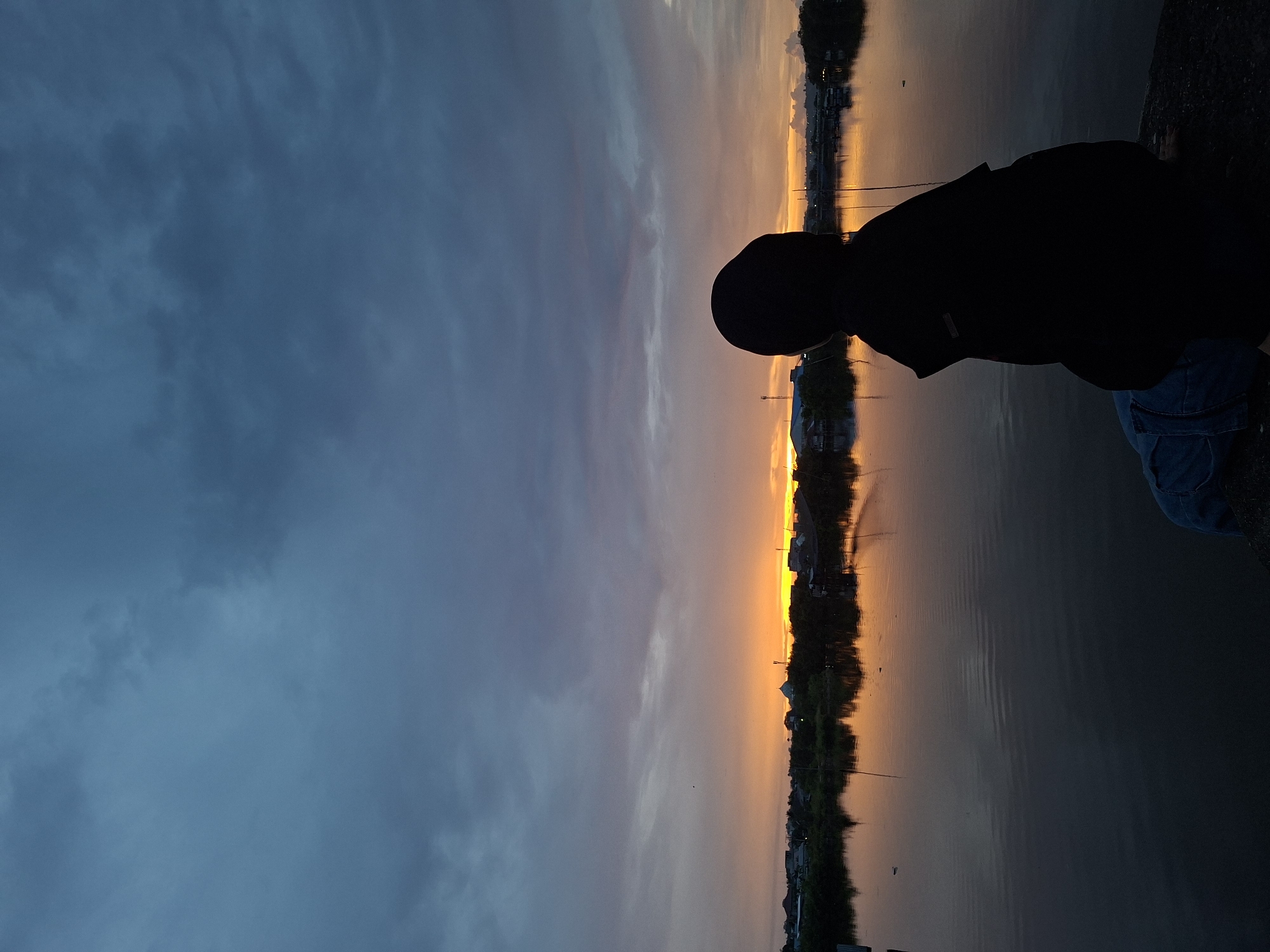 A long shot shows the silhouette of a person near a body of water under a partly cloudy sky during twilight in Lhokseumawe, Indonesia.
The person is dressed in dark clothing, including a hood and shirt, with a hint of blue jeans visible near their knees. They appear to be crouching or sitting near the edge of the water. The body of water reflects the colors of the sky and the buildings on the opposite bank.
The sky shows a gradient of colors, transitioning from a dark blue with clouds to an orange glow on the horizon. On the opposite bank, silhouettes of trees, buildings, and utility poles are visible. The image suggests a serene and tranquil setting by the waterfront during dusk or dawn in Lhokseumawe, Indonesia.