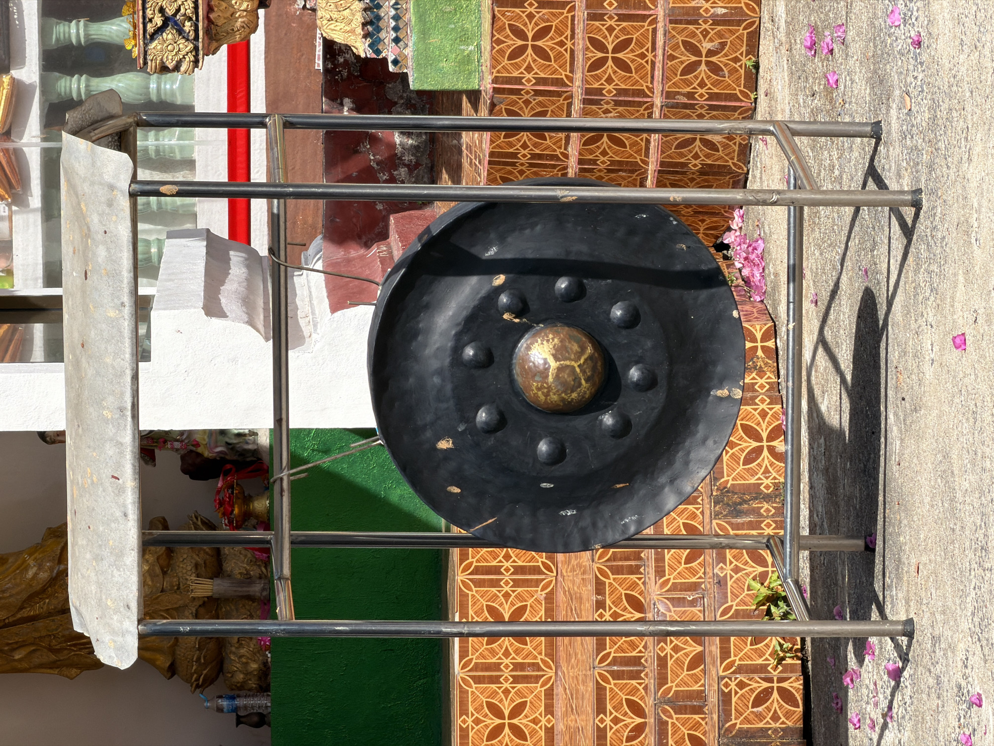 This is a full, eye-level, outdoor shot of a large, black gong mounted on a silver metal stand. The gong is circular with a raised, golden-brown center and several smaller circular bumps around the edge. The stand is a simple, utilitarian structure with a corrugated metal roof. Behind the gong and stand, there is a multi-tiered structure with decorative orange and brown tiles. To the left of the gong, a green wall is visible, and further left, a golden statue is partially obscured. To the right of the gong, there are steps and decorative elements of a shrine or temple. The ground in front of the stand is concrete, with scattered pink flower petals. The scene appears to be located in Baan Plai Laem, Thailand, likely within a temple or sacred site. There are no people visible in the image.