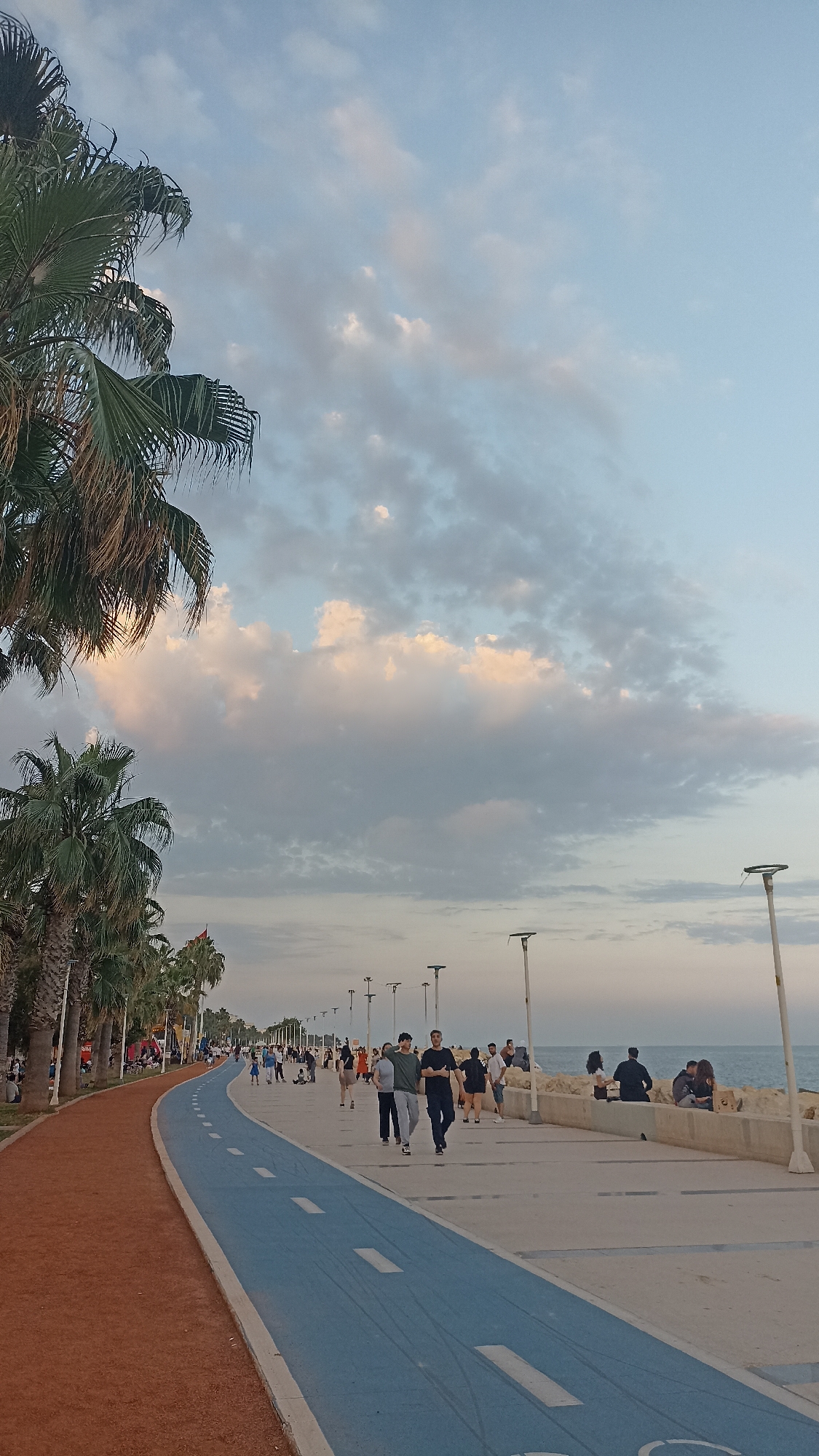 This image depicts a bustling coastal promenade under a partly cloudy sky. The scene is illuminated by natural light, suggesting either early morning or late afternoon, with some clouds showing hints of pink and orange.
On the left side of the frame, a dense line of tall palm trees with green fronds stretches into the distance, providing shade along the pathway. Adjacent to the palm trees is a wide, reddish-brown track, likely for jogging or running, which gently curves towards the left foreground. Immediately to its right is a blue track with white dashed lines, specifically marked for cycling, indicated by a faint bicycle symbol near the bottom right of the track. This blue track also follows the curve of the promenade.
To the right of the tracks, a spacious light-colored concrete pedestrian walkway is populated by numerous individuals and small groups. People are seen walking in various directions, some towards the viewer, others away. In the mid-ground, three men walk abreast towards the viewer, one in a light green shirt, one in a dark gray shirt, and one in a black shirt. Further along the promenade, people are observed strolling, interacting, and some are seated on a low concrete barrier that separates the walkway from the sea. Casual attire such as t-shirts, jeans, and shorts is prevalent among the pedestrians.
The right side of the image reveals a calm expanse of ocean, stretching to the horizon under the slightly hazy sky. A sturdy, light-colored concrete wall or barrier, interspersed with some natural rock formations, runs along the edge of the walkway, separating it from the water. A series of modern streetlights with distinctive flat, circular tops are evenly spaced along the promenade, primarily on the sea-facing side. In the mid-distance on the left, amidst the palm trees, a red flag is visible on a pole. The overall environment appears recreational and vibrant.