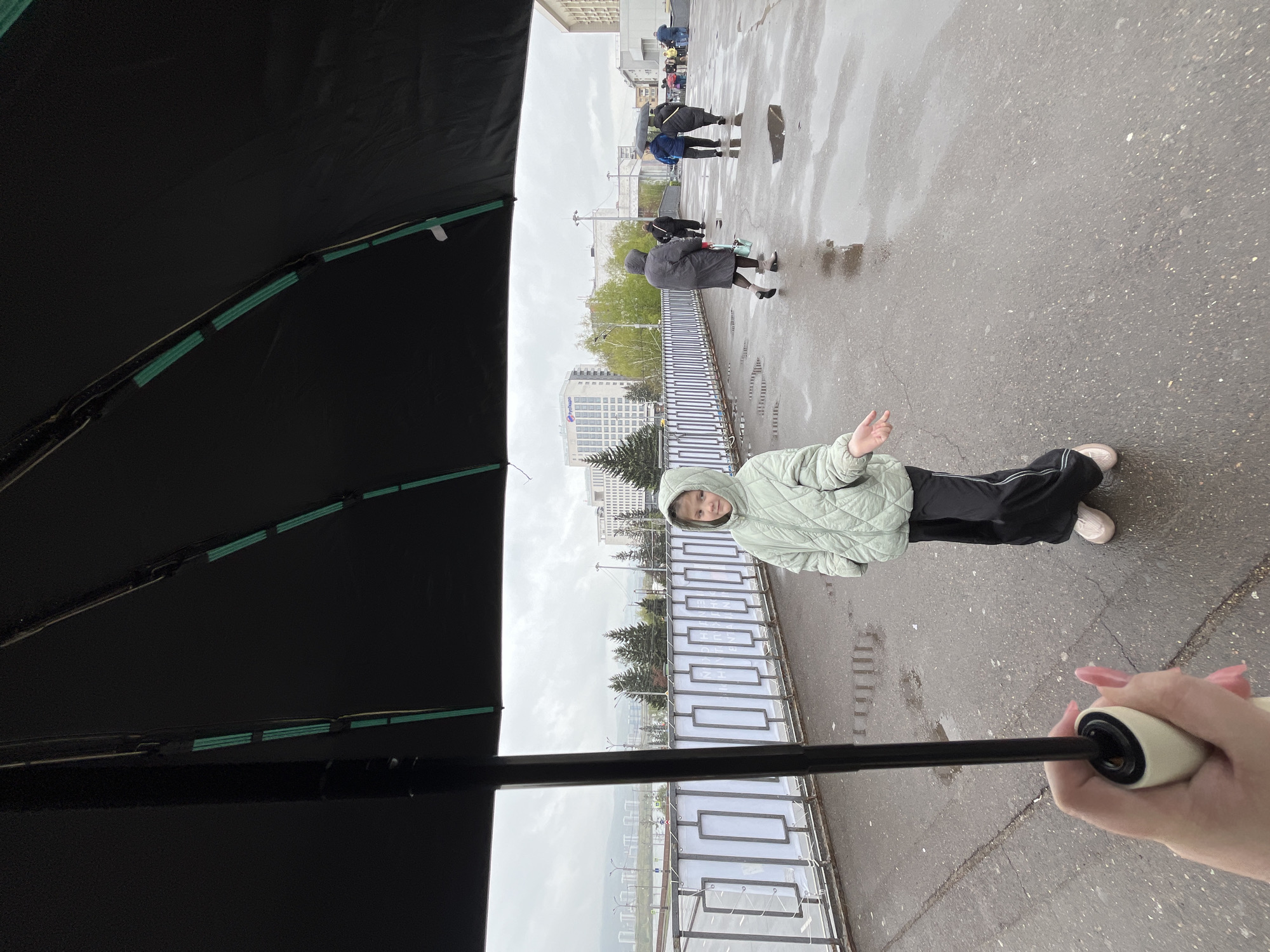 Here is a detailed description of the image:
**Scene Overview:**
The image is taken from the perspective of someone holding an umbrella on a rainy day in what appears to be an urban area. The composition is somewhat obscured by the umbrella itself, which dominates the foreground. The person holding the umbrella is observing a young person standing on a bridge or elevated walkway and interacting with them. Further in the background, other people can be seen walking in the same direction.
**People:**
1. A person is holding the umbrella. Only their hand is visible.
2. A young person stands on the walkway, wearing a light green quilted jacket with a hood and dark pants. They are making a gesture with their hand.
3. Several other people are walking in the distance, some also carrying umbrellas.
**Objects:**
1. A black umbrella is prominent in the foreground, partially obscuring the scene.
2. A metal railing borders the walkway or bridge.
3. Buildings are visible in the background.
4. Road with puddles and pedestrians is visible in the background.
**Environment:**
The weather is rainy, indicated by the umbrella being used and the wet surfaces. The scene is set in an urban environment, with buildings, roads, and trees in the background.
**Location:**
This is Krasnoyarsk, Russia. The background features the cityscape of Krasnoyarsk, characterized by the presence of modern buildings.