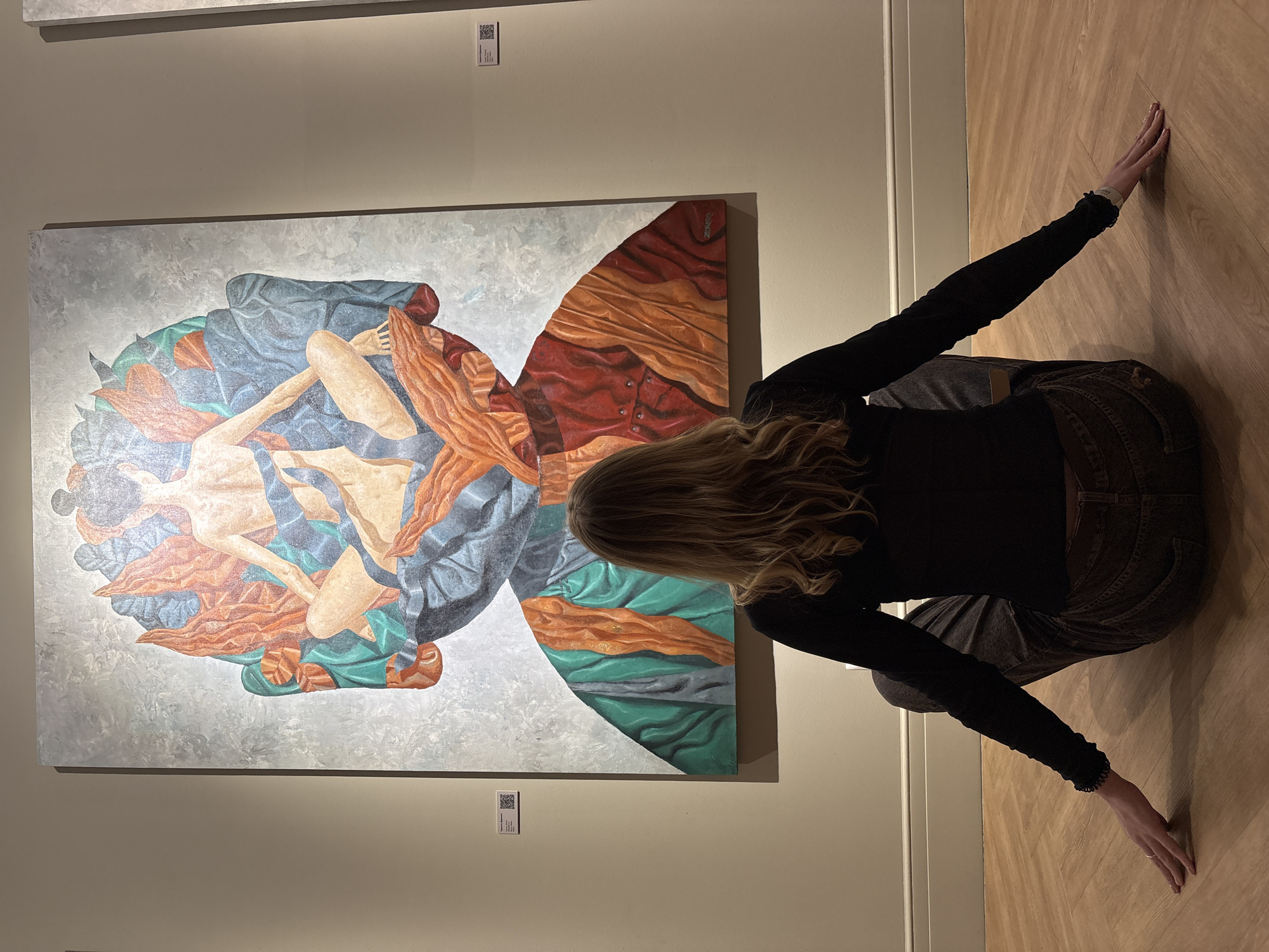 A young woman with long blonde hair kneels on a light brown wooden floor in front of a large abstract painting. She faces away from the camera, her arms outstretched so that her hands touch the floor on either side of her. She wears a black long-sleeved shirt and dark-colored jeans. The painting, which is predominantly gray, blue, red, orange, and green, features intertwined figures. The scene is likely inside a museum or art gallery with light beige walls. The location is not verifiable as Irkutsk, Russia.
