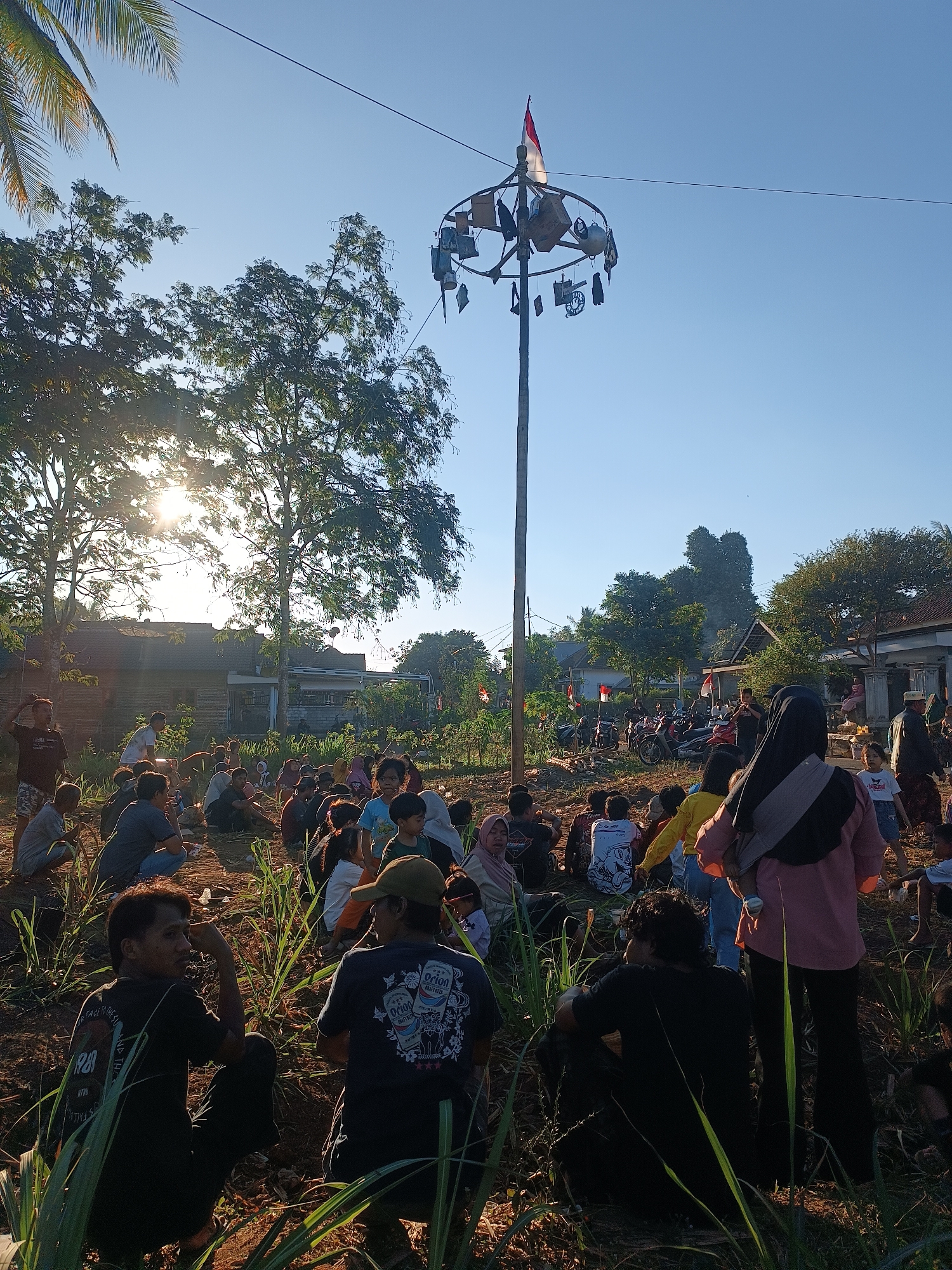 In Balearjo, Indonesia, a community gathers outdoors under a clear blue sky, the sun casting long shadows and glinting through lush green trees. A tall pole, adorned with various objects and topped with an Indonesian flag, stands prominently in the center of the scene. A crowd of people, dressed in casual attire, are seated on the ground, some facing the pole, others interacting with each other. Among them are children and adults, suggesting a family-friendly event. The ground is uneven and appears to be covered in dry leaves and sparse vegetation. In the background, residential buildings and parked motorcycles are visible, indicating a semi-rural setting. The overall atmosphere seems to be one of communal gathering and celebration.