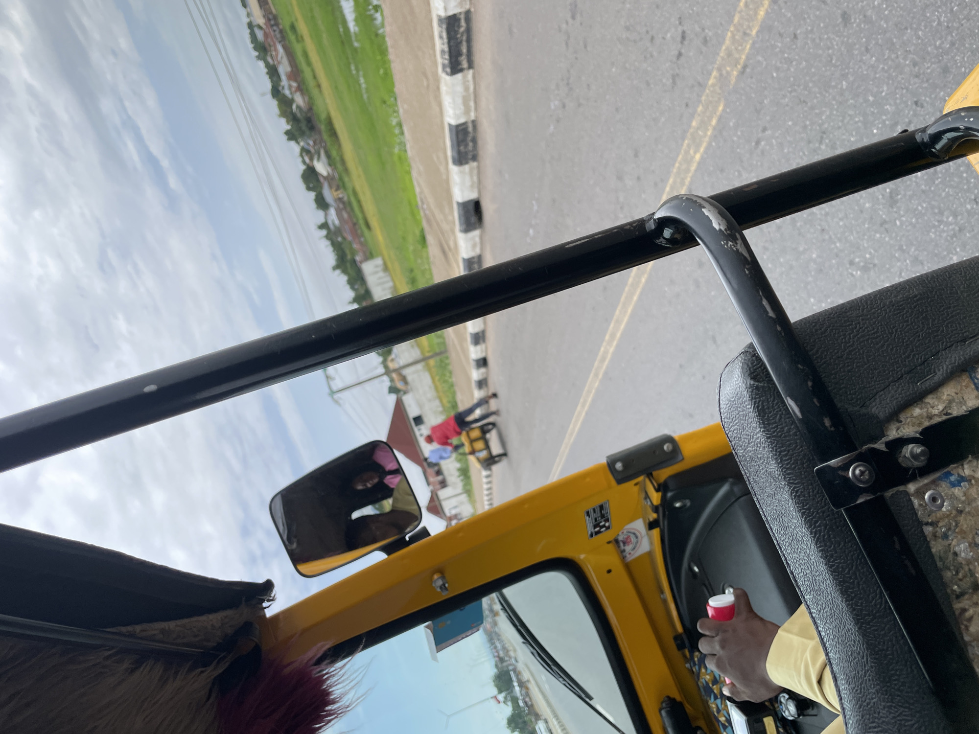 The image shows a driver's perspective from inside a yellow motorized rickshaw, commonly known as an "okada" or "keke," in Jalingo, Nigeria. The driver's hand, wearing a light yellow long-sleeved shirt, is visible on the steering column, holding a pink and white object. Reflected in the side mirror are two individuals, a man in a light-colored shirt and a woman in a pink top. Outside the rickshaw, on the right side of the road, a person in a red shirt is pushing a yellow handcart. The road has a black and white striped curb. In the background, there is a green grassy area, possibly a field or marsh, with buildings and trees in the distance. The sky is overcast with some visible power lines. The scene appears to be on a street within Jalingo.