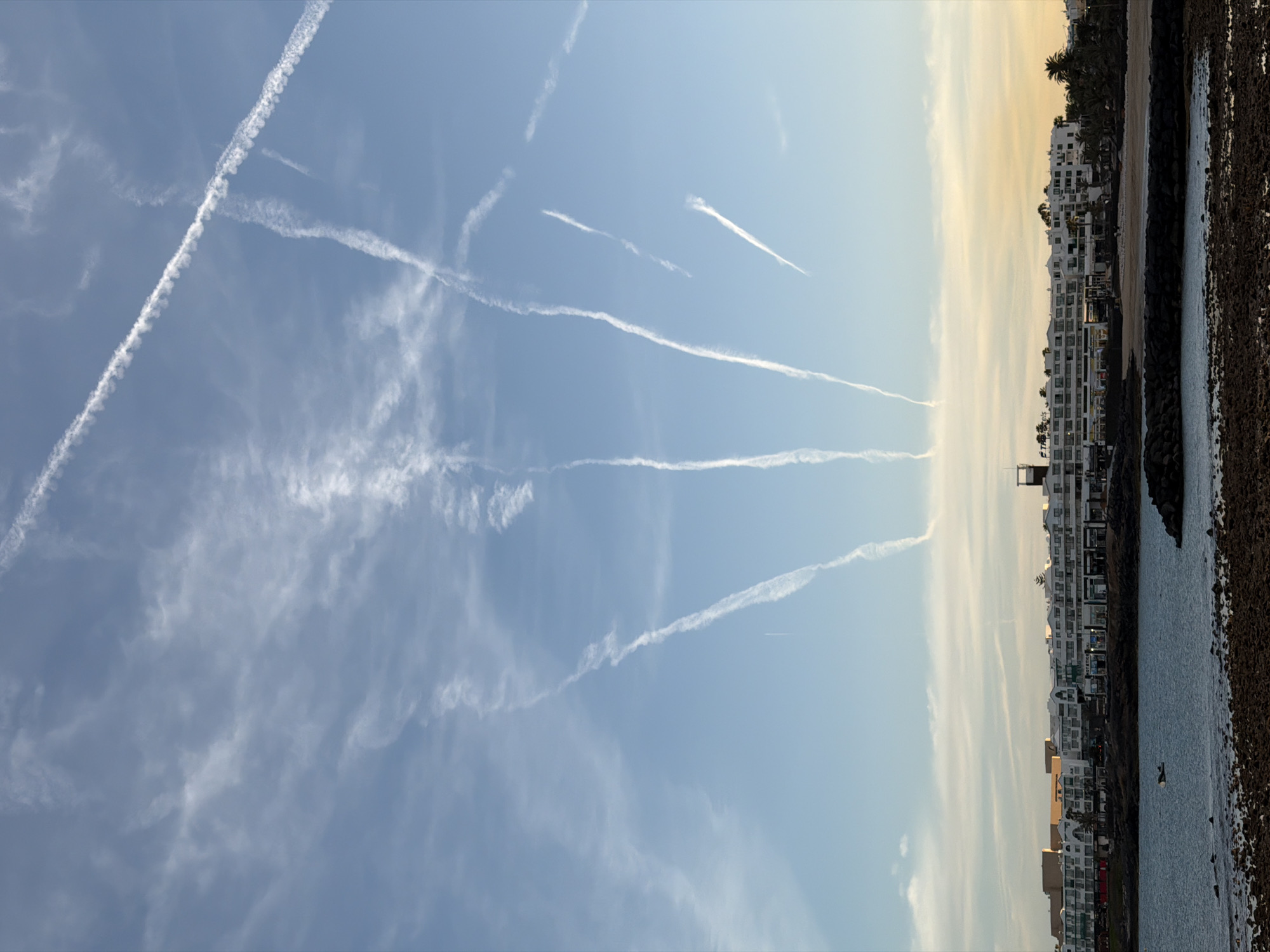 The image displays a vibrant coastal scene under a sky crisscrossed with numerous contrails, suggesting a busy air traffic route or specific atmospheric conditions. The sky, predominantly a clear light blue, occupies the upper two-thirds of the frame. It features multiple prominent white contrails; some are sharp and linear, while others are broader and more diffused, indicating different stages of dissipation. One long, distinct contrail stretches diagonally from the upper left towards the lower right. Wispy, high-altitude clouds are also present, particularly denser in the lower-left corner of the sky.
Along the horizon, a row of modern, white multi-story buildings with many windows and balconies stretches across the lower third of the image. Behind some of these structures, dark green palm trees are visible, hinting at a tropical or resort setting. The lighting suggests either late afternoon or early morning, with a soft, warm yellow-orange glow tinting the sky directly above the buildings and reflecting on their facades.
Adjacent to the buildings, on the right side of the frame, is a calm body of water, likely the ocean or a bay. A dark, rugged rocky shoreline or breakwater separates the buildings from the water. A small, dark object, possibly a person or a small boat, is discernible in the water in the distance. No visible text is present. The overall scene depicts a sunny day in a coastal urban area, consistent with Teguise, Spain.