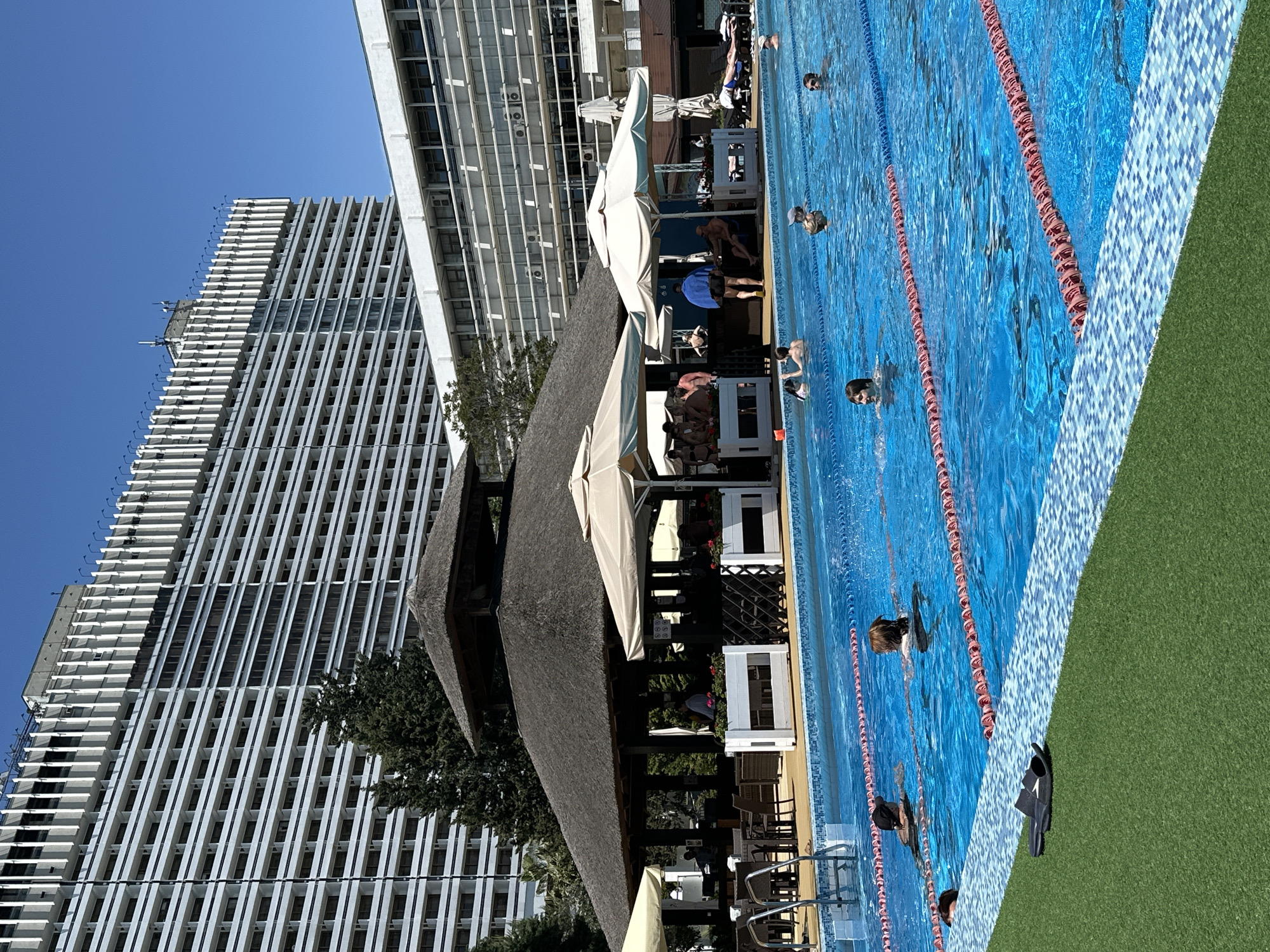 The image shows a swimming pool next to a building in Sochi, Russia. Several people are in the pool. There are lane markers in the pool. A building is in the background. There are several tables and chairs in the foreground next to the pool. There is artificial grass next to the pool.