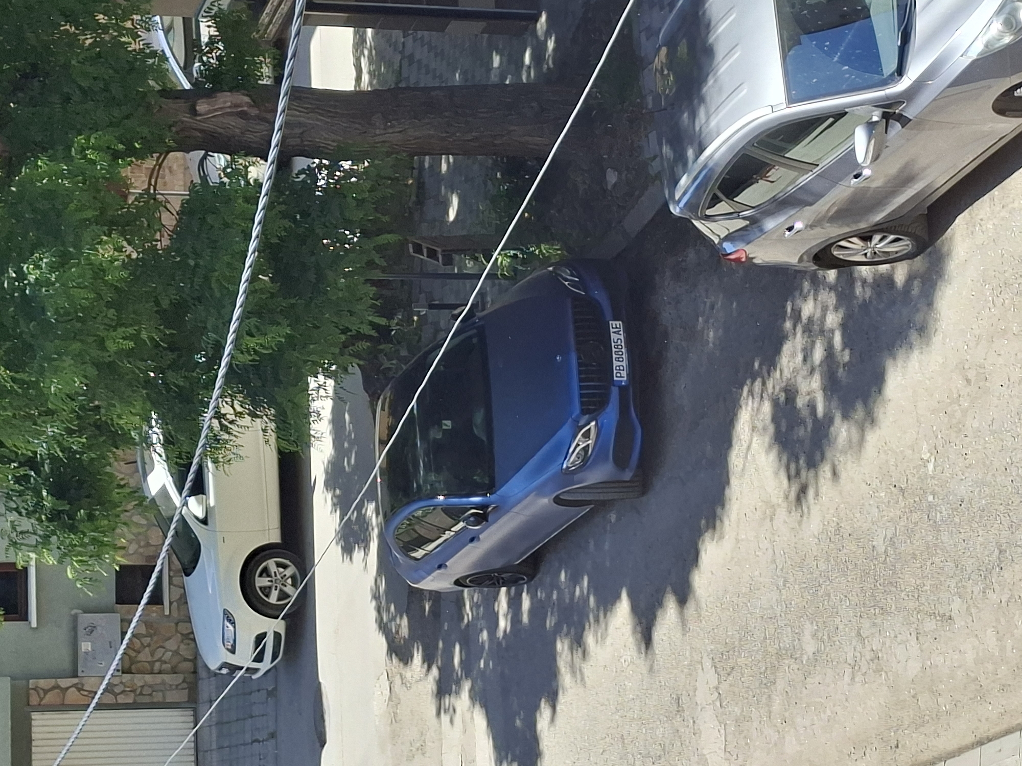 The image depicts a street scene in Plovdiv, Bulgaria, featuring three parked cars. A silver sedan is positioned on the right, a blue coupe in the center, and a white SUV on the left. The blue coupe has a visible license plate reading "PB 8885 AE." The street is shaded by trees, casting dappled shadows on the pavement. Overhead power lines are visible. The environment appears to be a residential area with buildings and landscaping in the background. No people are visible.