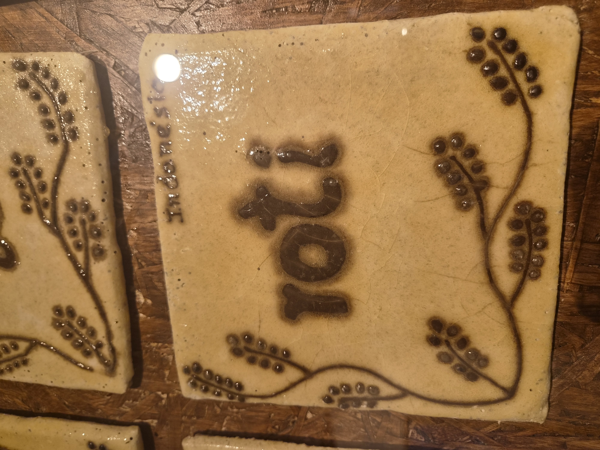 This is a close-up, eye-level, indoor shot of a square, light-brown tile with a dark brown floral design and the word "roti" stamped on it. The tile is positioned on a dark wooden surface. The word "roti" is in the center of the tile, and the floral design consists of thin, branching lines with clusters of small dark dots, resembling berries or seeds. There are also faint markings above the word "roti" that appear to be text, possibly "Indonesia". The tile has a slightly glossy finish and some minor imperfections and cracks. There are no people or animals visible in the image. The lighting creates a reflection on the surface of the tile, obscuring some of the details. The image does not provide any information about the city of Seia, Portugal, as it is not depicted in the scene.