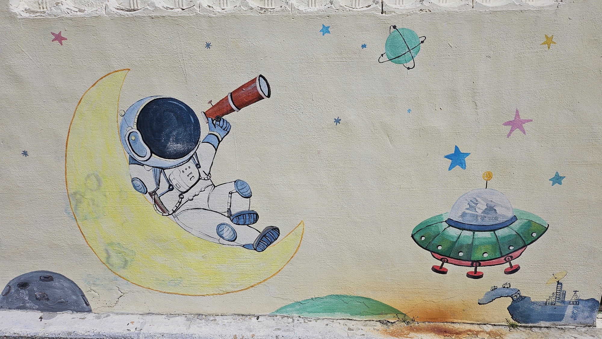 The image is a photograph of a mural, located in Ulsan, South Korea. The mural depicts an astronaut sitting on a crescent moon, holding a red telescope. To the right, there is a green flying saucer with a transparent dome, containing two figures. Scattered around are colorful stars, and a green-blue planet. At the bottom of the mural, there are gray, green, and blue landmasses painted. The mural is on a pale beige wall, with some damage near the base.