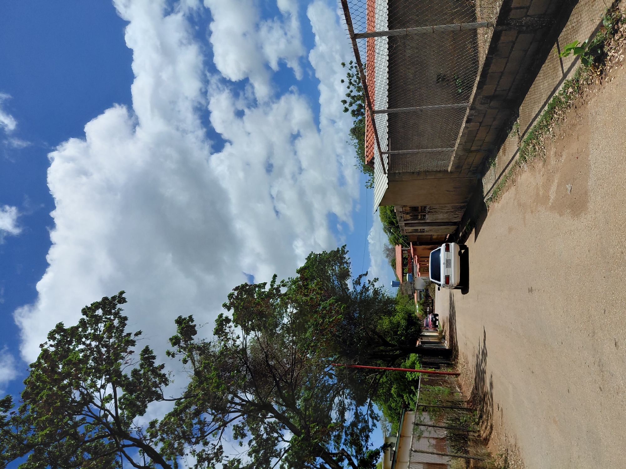 A dirt road winds through a residential area in Santa Ana, Venezuela. On the left side of the road, a chain-link fence encloses a property with a building behind it. A large tree with lush green foliage casts a shadow over the road. Further down the road, two cars are parked, one white sedan and a darker colored SUV. On the right side of the road, another chain-link fence with a concrete base separates the road from a property. Behind the fence, a building with a red-tiled roof is visible. The sky above is a vibrant blue, dotted with fluffy white clouds. The overall scene depicts a sunny day in a quiet neighborhood.