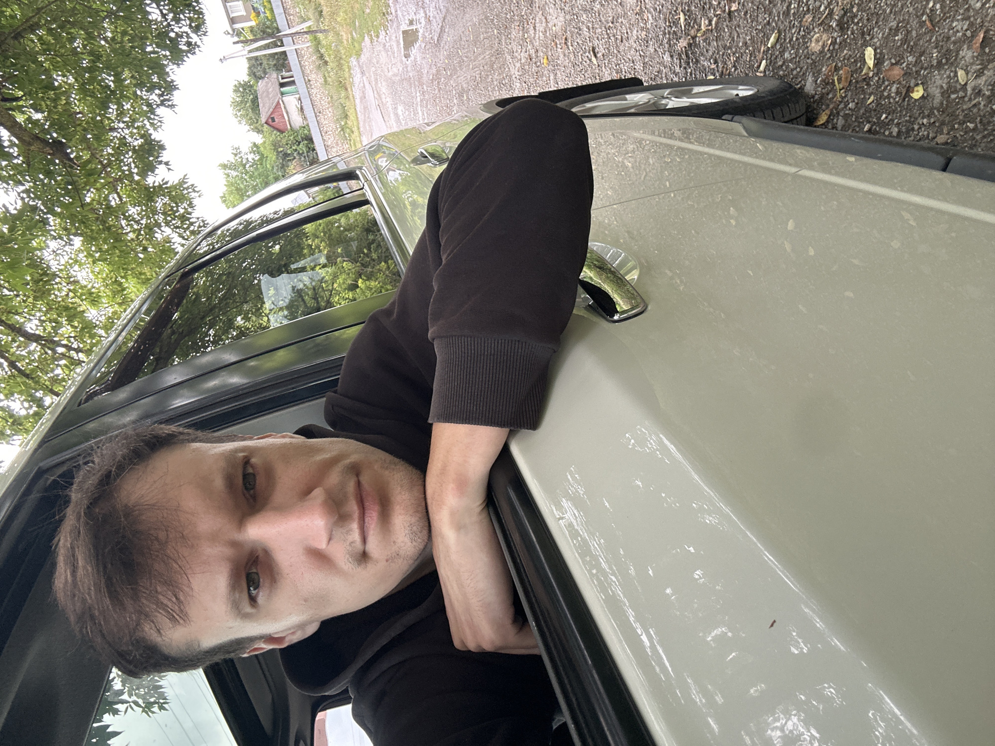 A man is leaning out of a car window, resting his arms on the door frame and looking at the camera. The car is parked on a gravel road, with trees and houses visible in the background. The environment appears to be rural. The man is wearing a dark hooded sweatshirt. The car is light-colored.
