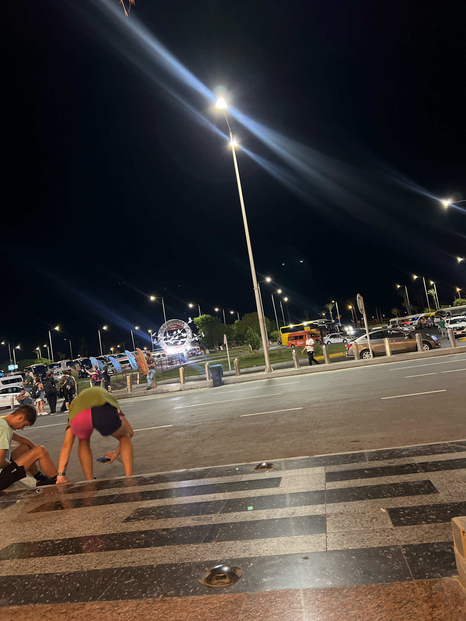 The image shows a nighttime scene in the city of Sharm El Sheikh, Egypt, likely in an area with shops and entertainment. In the foreground, two young men are sitting on a tiered stone structure. One is wearing a light green t-shirt and dark shorts, with his legs crossed. The other, wearing a lime green and pink athletic top and dark shorts, is bending over, looking at his phone. Behind them, a street with parked cars and buses is visible. In the midground, a circular illuminated sign with the word "SOHO" can be seen, surrounded by palm trees and streetlights. People are gathered around the sign and walking along the street. The sky is dark, with prominent light streaks from streetlights and possibly fireworks or other aerial lights. The overall impression is of a lively evening atmosphere in a tourist or entertainment district.