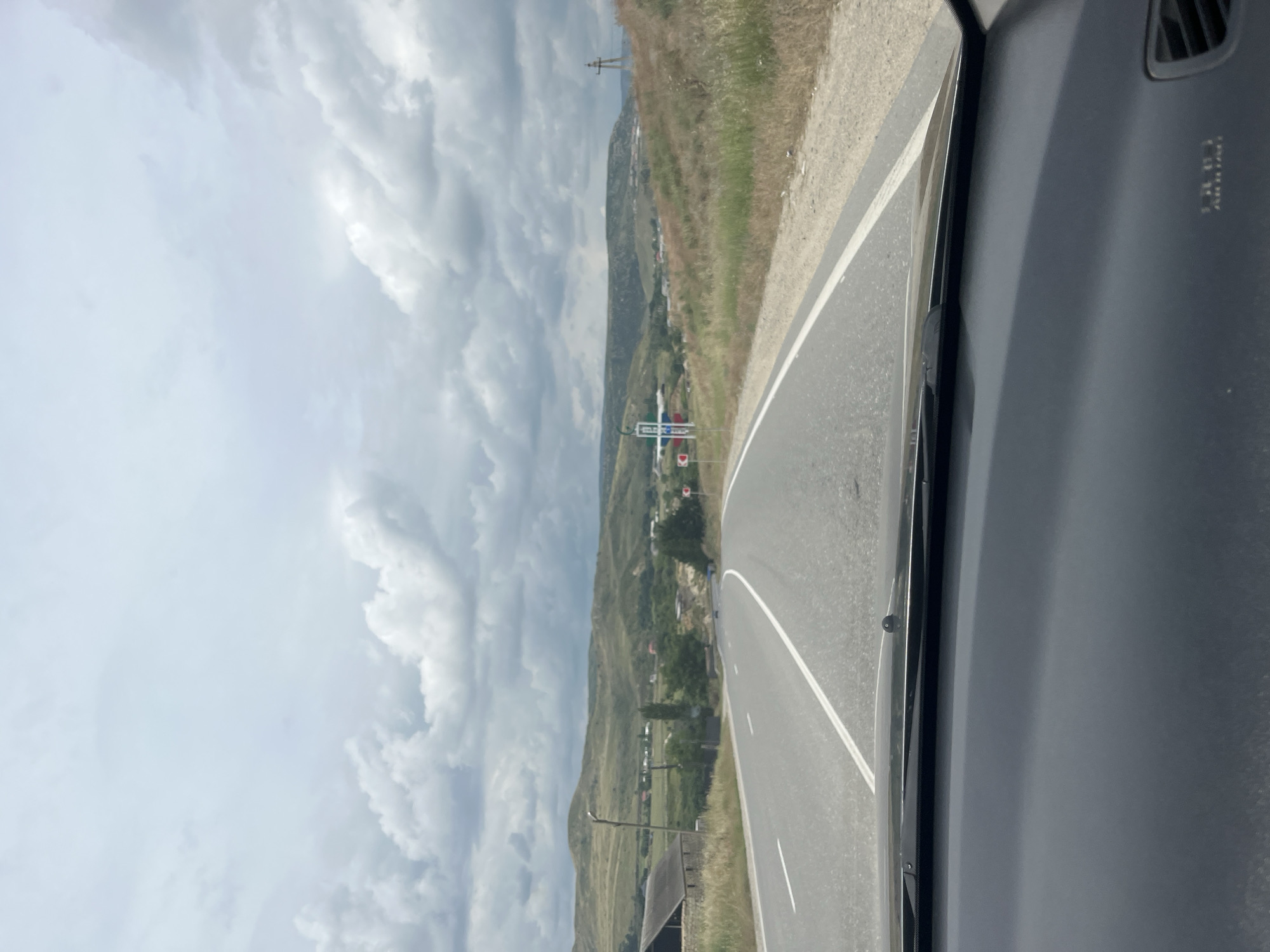 The image captures a view from inside a vehicle traveling on a paved road. The road is asphalt with distinct white lane markings. On the left side of the road, there is a building with a dark roof and stone walls, and a tall, thin structure resembling a lamppost or utility pole. The right side of the road features dry, yellowish grass and a gentle slope leading up to a grassy hillside dotted with bushes and small trees. In the distance, rolling green hills form the backdrop, with a smattering of houses and buildings visible in the valley. The sky is overcast with a layer of thick, gray clouds, suggesting a cloudy or potentially rainy day. There are signs visible on the side of the road, including what appears to be a directional sign with a red and white symbol and another sign with a blue and red design. A tall, cross-shaped structure, possibly a power line tower, stands on the right-hand hillside. The foreground shows the dark dashboard of the vehicle. The scene appears to be a rural or semi-rural area, likely in the vicinity of Karabudakhkent, Russia, given the context of the request.