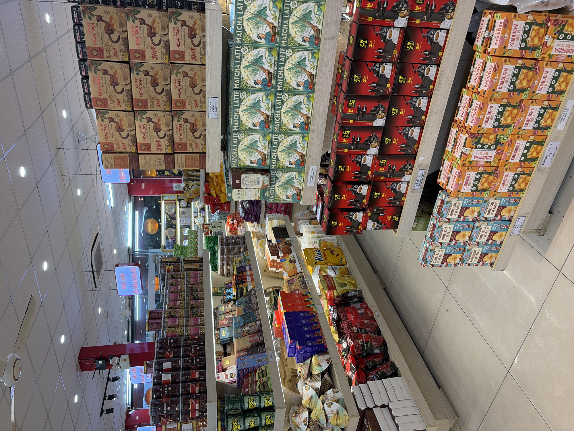 The image depicts the interior of a well-lit retail store, likely a supermarket or a specialty food shop, in Da Nang, Vietnam. The shelves are stocked with a variety of food and beverage products. Prominently displayed are boxes of coffee, matcha latte, and kombucha, arranged in neat stacks. There are also shelves filled with jars of what appear to be dried fruits or snacks, as well as packaged goods like biscuits and boxes of other food items. In the background, there are illuminated signs, one of which reads "BÁC SẢN". A ceiling fan is visible in the upper left portion of the image, and the floor is tiled with light-colored square tiles. The overall impression is a clean and organized retail environment offering a selection of consumer goods.