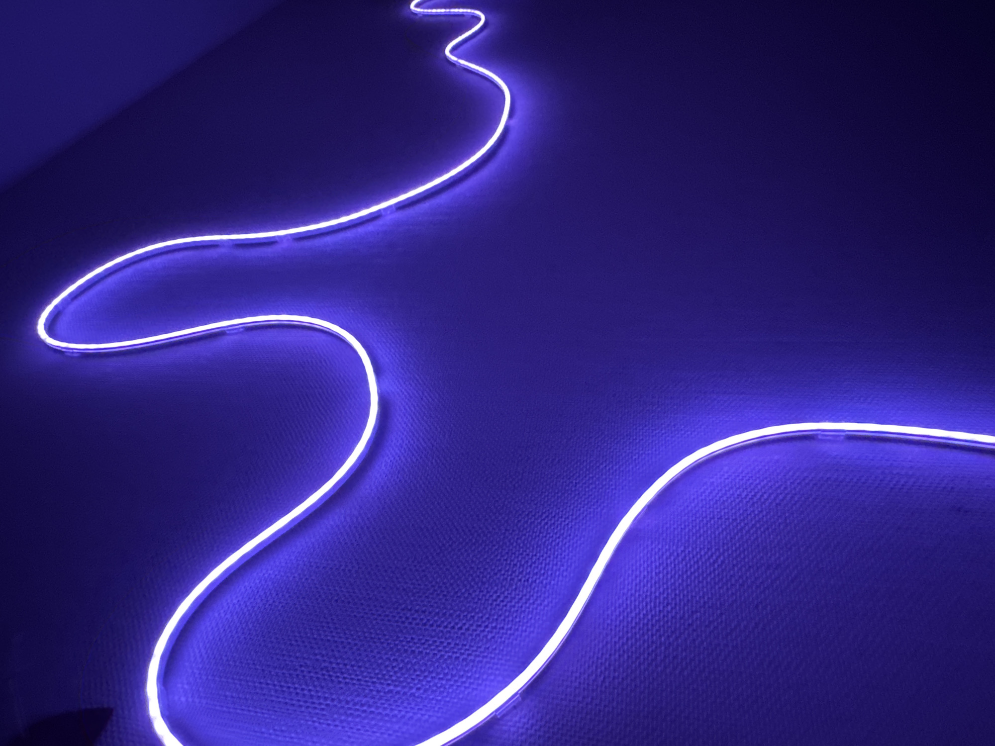 The image displays a flexible, white-glowing LED strip light affixed to a textured wall, creating an undulating, wave-like pattern. The wall itself is a deep purple or indigo hue, exhibiting a subtle woven or stippled texture, particularly noticeable where illuminated by the light strip. The ambient lighting appears low, with the glowing strip serving as the primary light source, casting a soft luminescence onto the immediate wall surface. No individuals, additional objects, or discernible actions are present in the frame. The scene is located in the city of Perm, Russia.