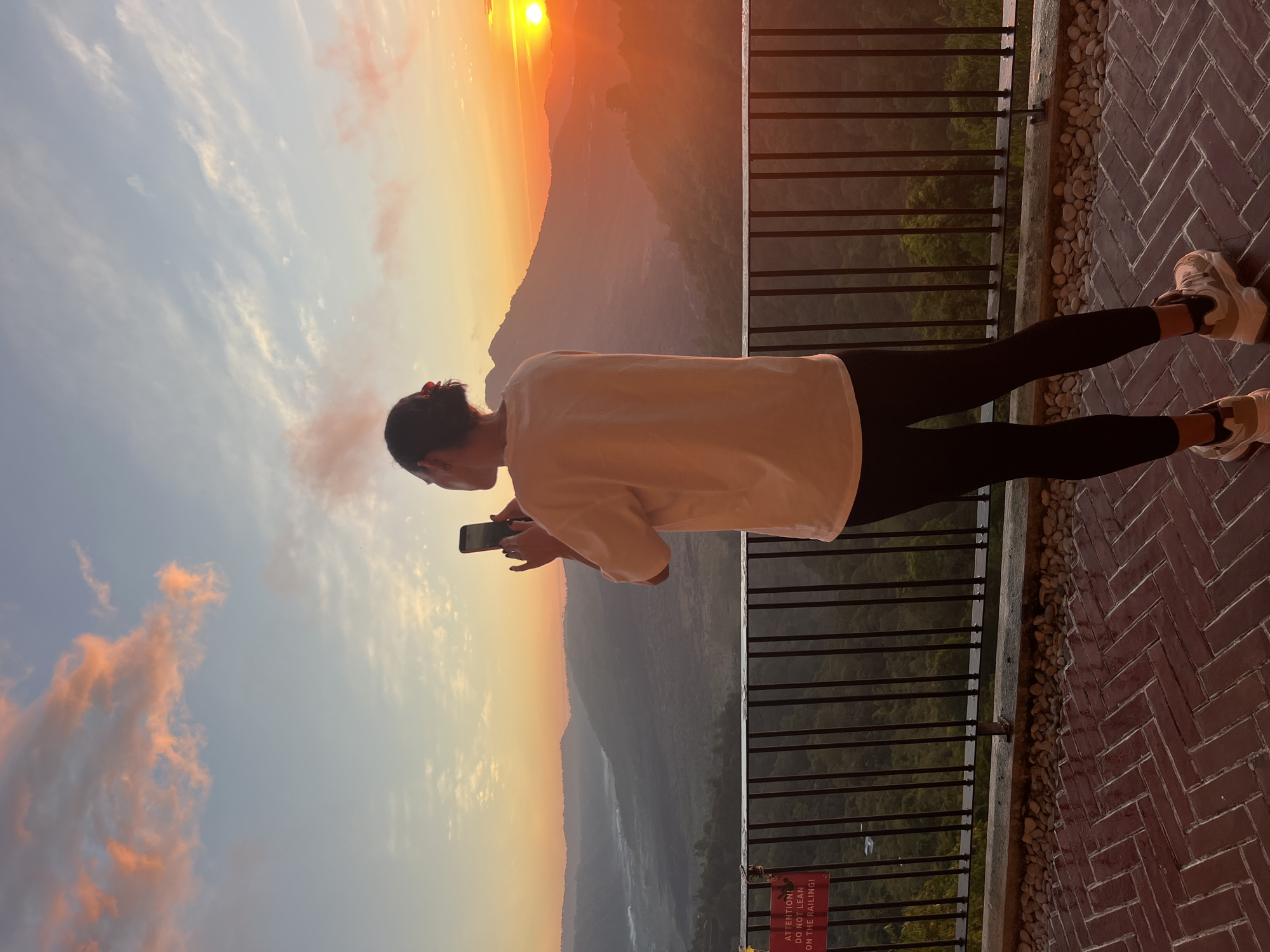 The image depicts a person standing on a tiled walkway, holding a smartphone and seemingly taking a picture. They are wearing a white t-shirt, black leggings, and athletic shoes. Behind the person is a railing, beyond which lies a scenic overlook of a valley with mountains in the distance, bathed in the warm light of a sunset. A sign attached to the railing reads "ATTENTION! DO NOT LEAN ON THE RAILING!". The sky is painted with hues of orange, pink, and blue from the setting sun, with scattered clouds. The environment appears to be a viewpoint in Kintamani, Indonesia, known for its stunning views of Mount Batur and Lake Batur. The overall scene captures a moment of appreciating the natural beauty of the location during sunset.